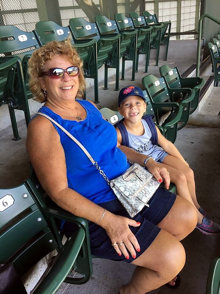 Smiling woman in blue top and sunglasses sits with a young girl in a stadium, green empty seats behind them.