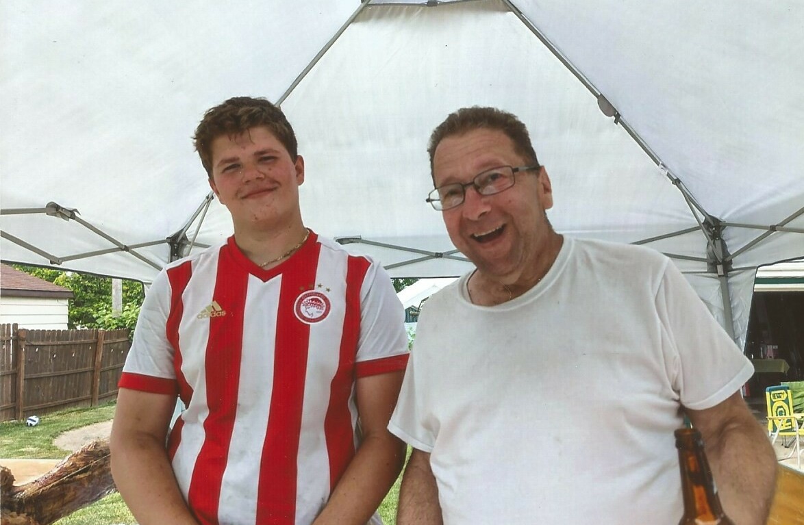 Two men pose together under a white canopy in a backyard; one wears a red‑and‑white striped soccer jersey, the other a plain white shirt, both smiling.