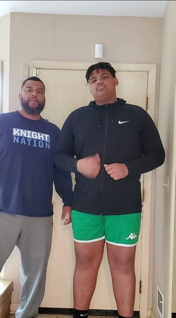 Two men posing for a photo indoors, one wearing a navy Knight Nation shirt and gray pants, the other in a black zip hoodie and green shorts.