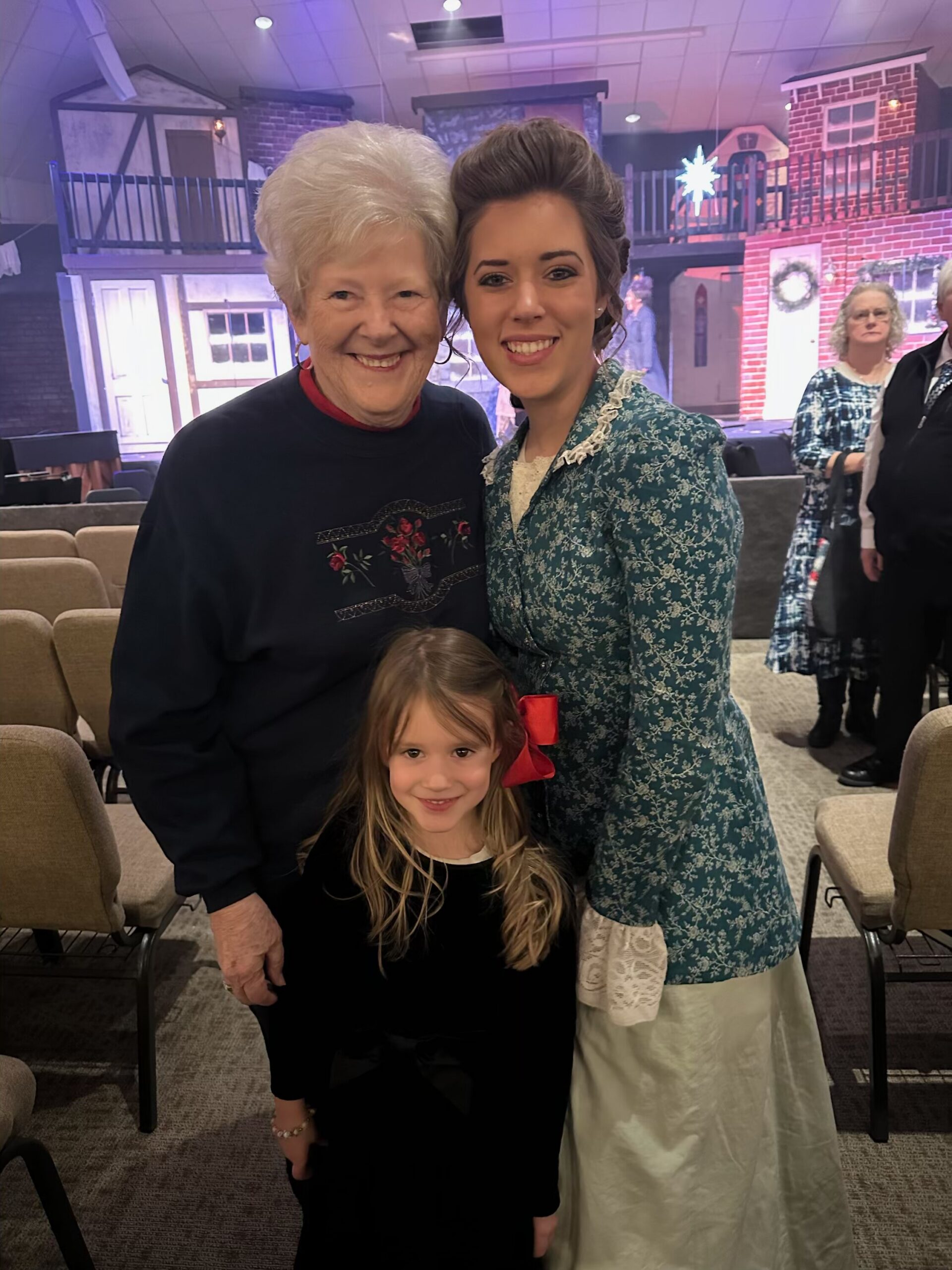 Three generations posing together in a theater lobby: grandmother, young woman in a teal patterned dress, and a small girl in black, smiling at the camera with a stage backdrop behind them.