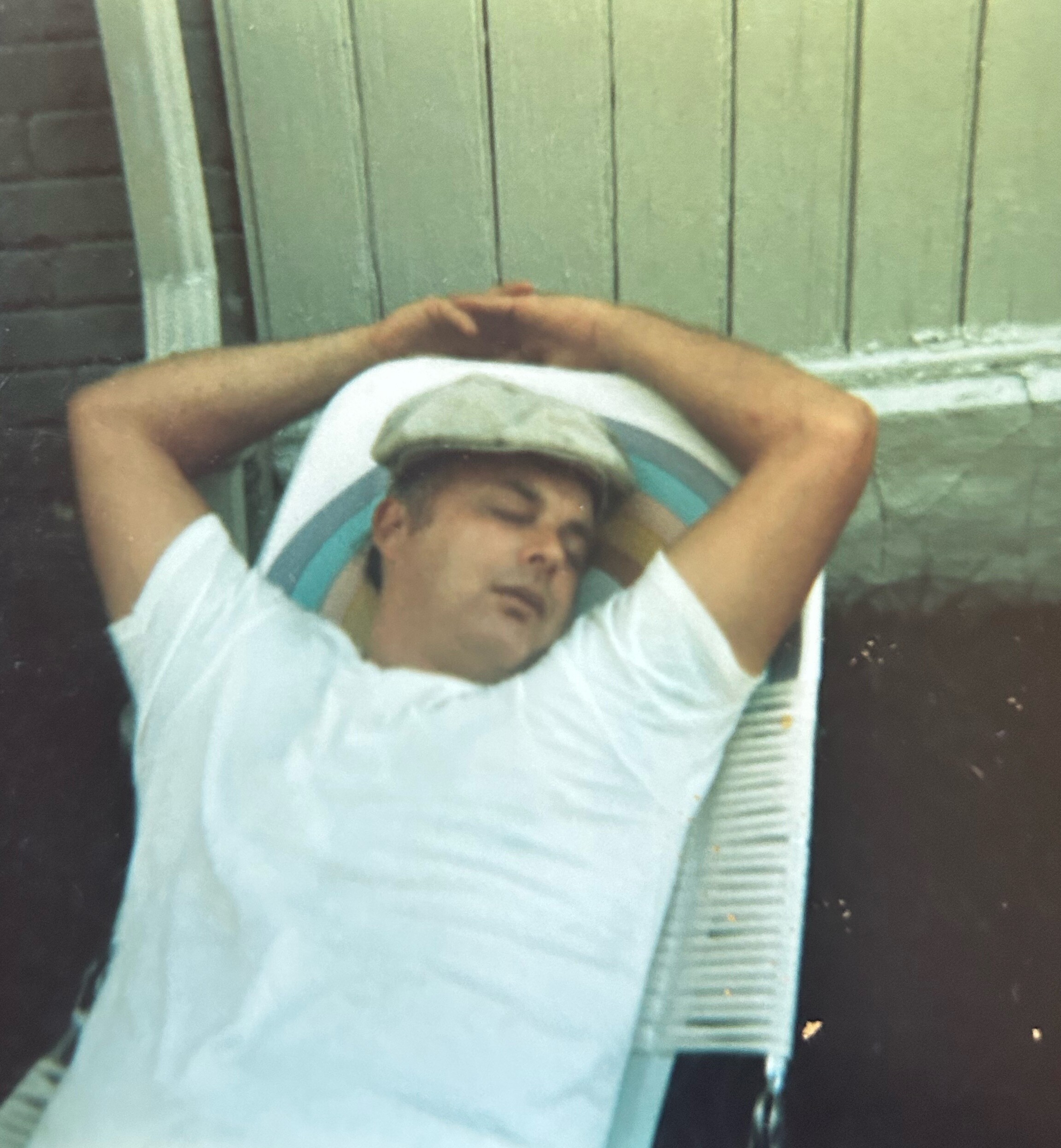 Man resting with arms folded behind his head in a striped chair, wearing a light cap outdoors near a green fence.