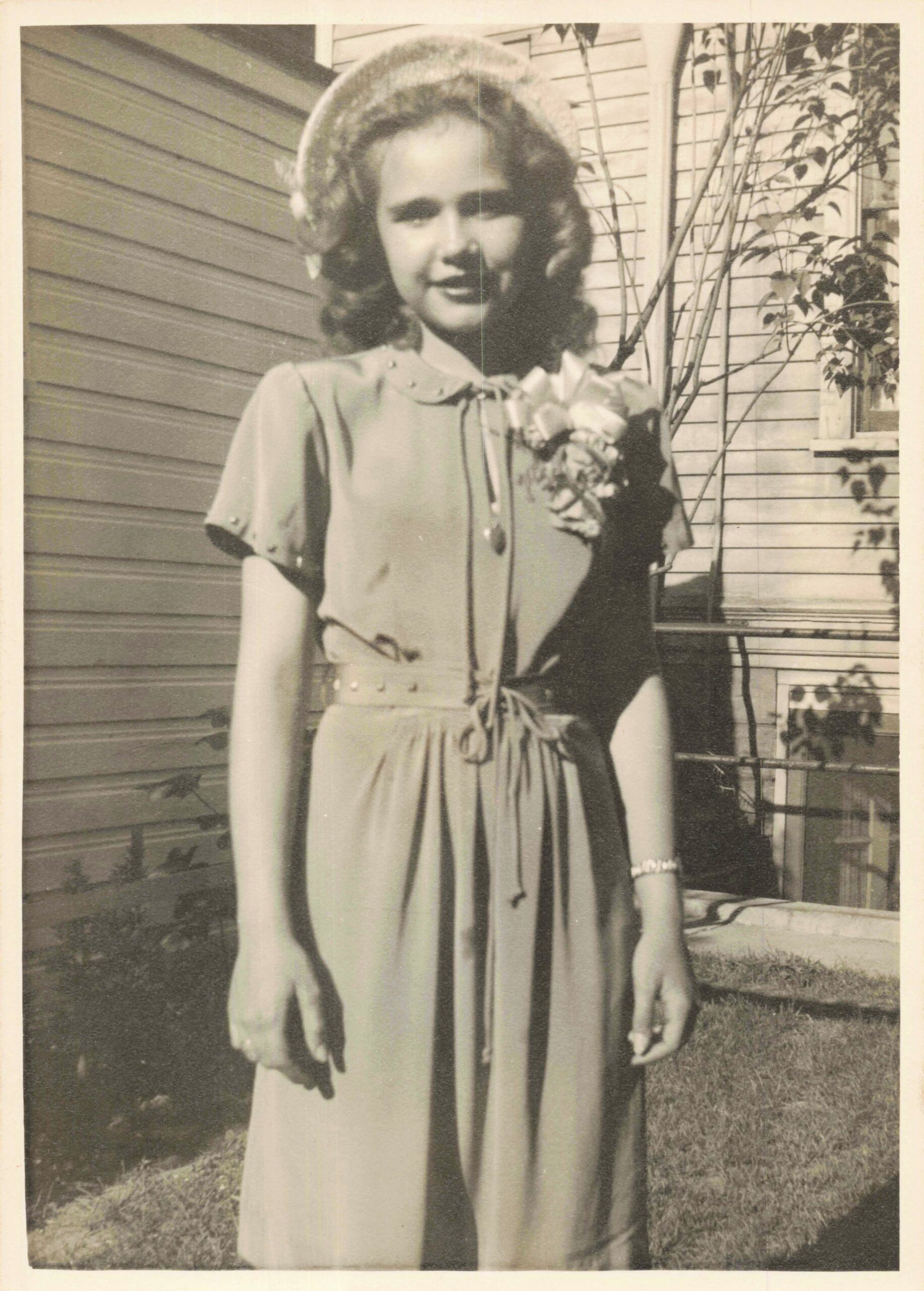 Young woman in a belted dress with a floral corsage, standing outside a house with siding and climbing plants.