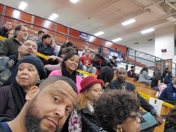 A diverse group of people sits in gym bleachers, with a man taking a selfie in the foreground and others smiling behind him at a community event.