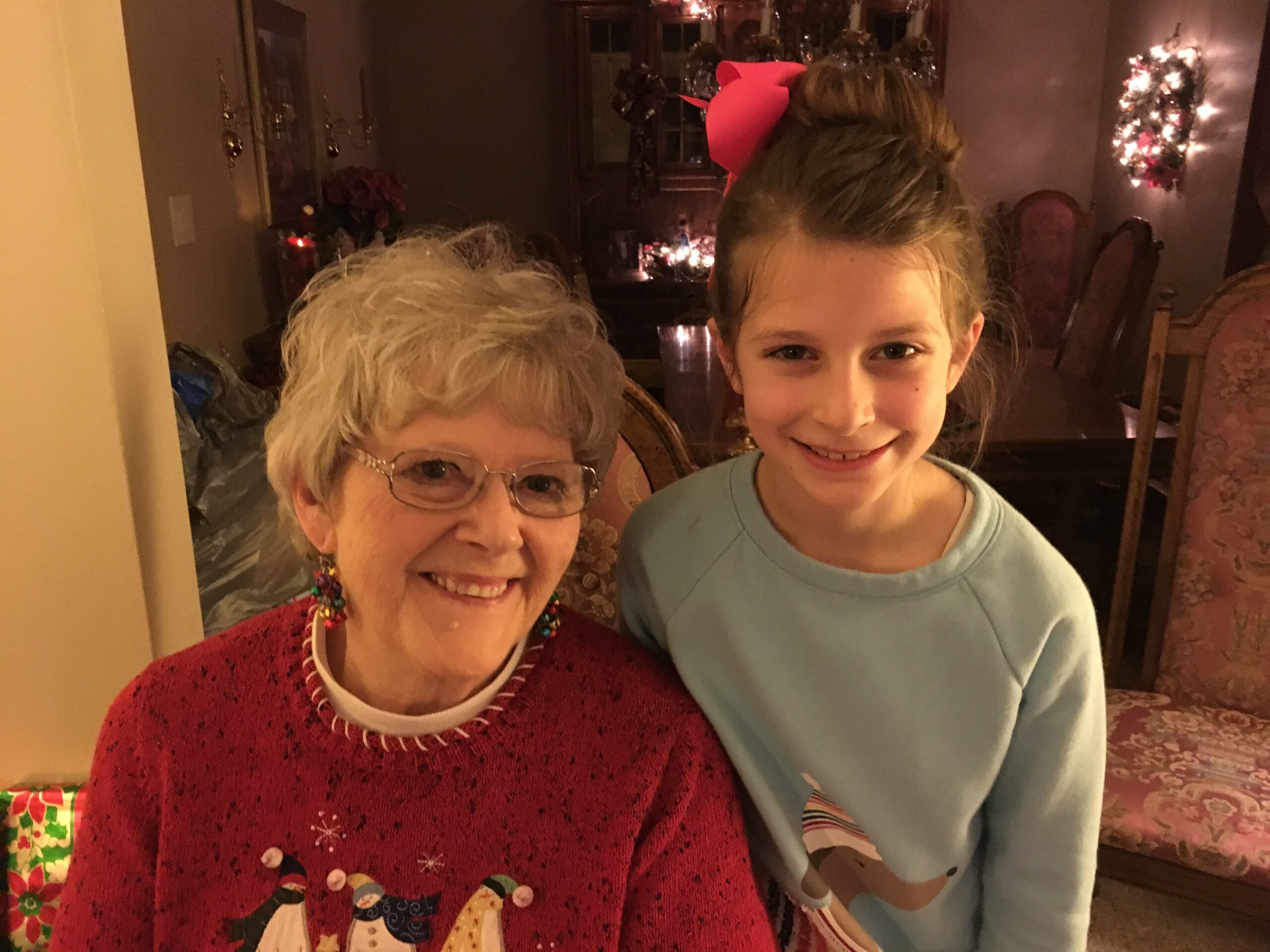 Smiling elderly woman in a red Christmas sweater beside a smiling girl with a red bow in her hair, holiday decorations in the background.