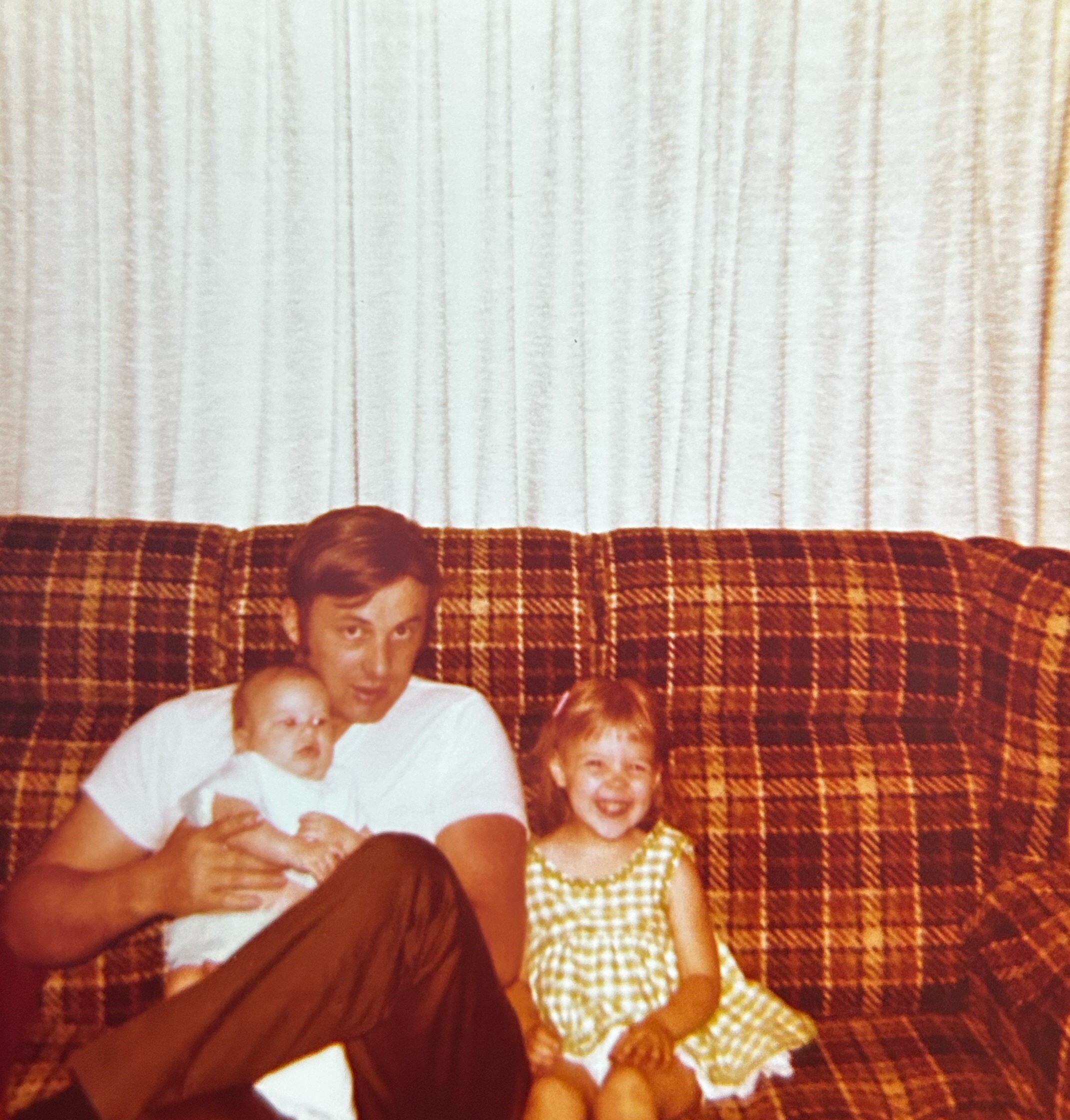 Man seated on a brown plaid couch holds a baby while a young girl in a checkered dress stands beside him in a living room setting.