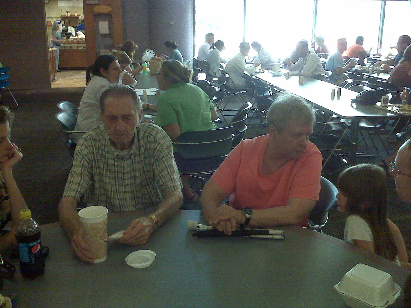 Two older adults sit at a table in a busy cafeteria, with cups and a bottle nearby, as others eat in the background.