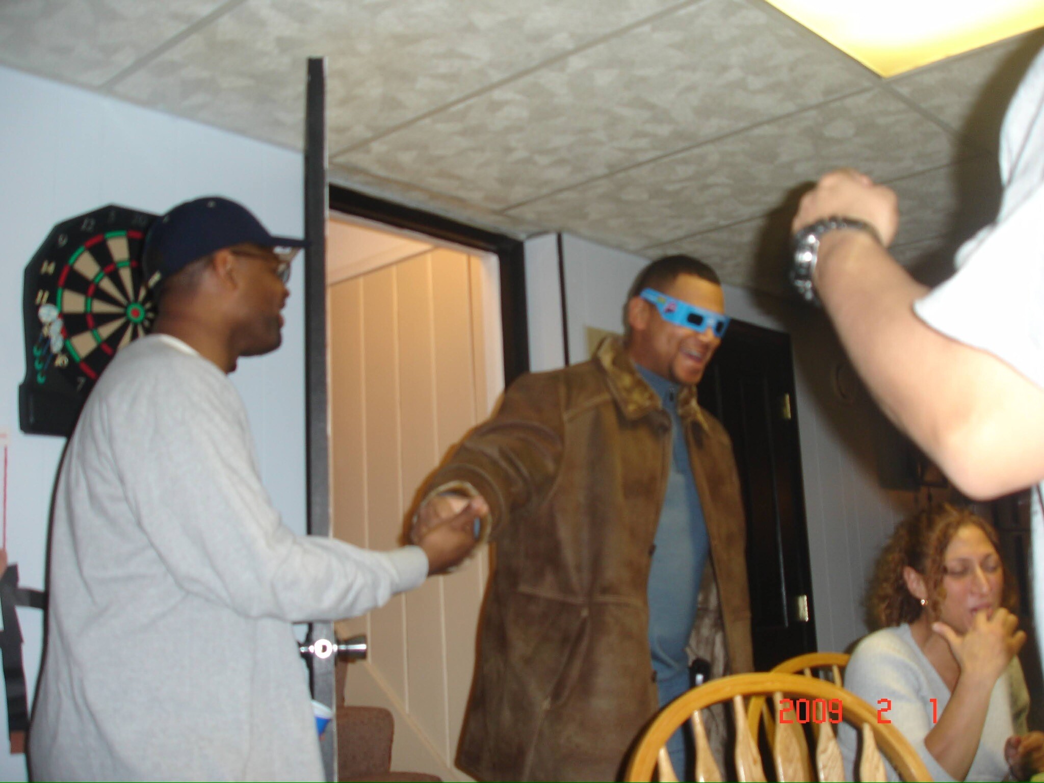 Indoor social gathering with several people standing and sitting; a dartboard on the wall and a person wearing novelty blue 3D glasses in the center.