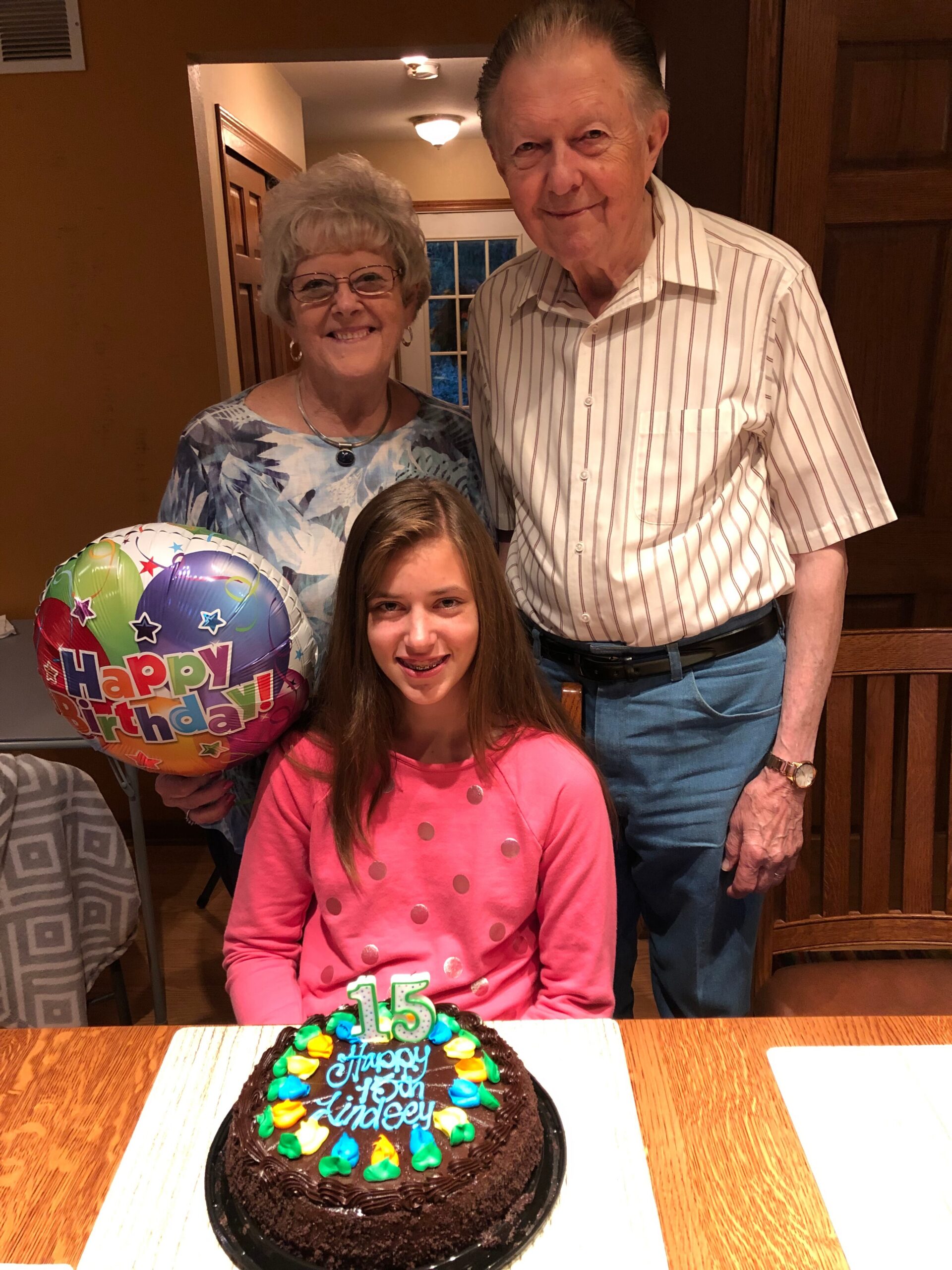 Young girl in pink sits at a table with a chocolate birthday cake, flanked by smiling grandparents and a balloon in the background.
