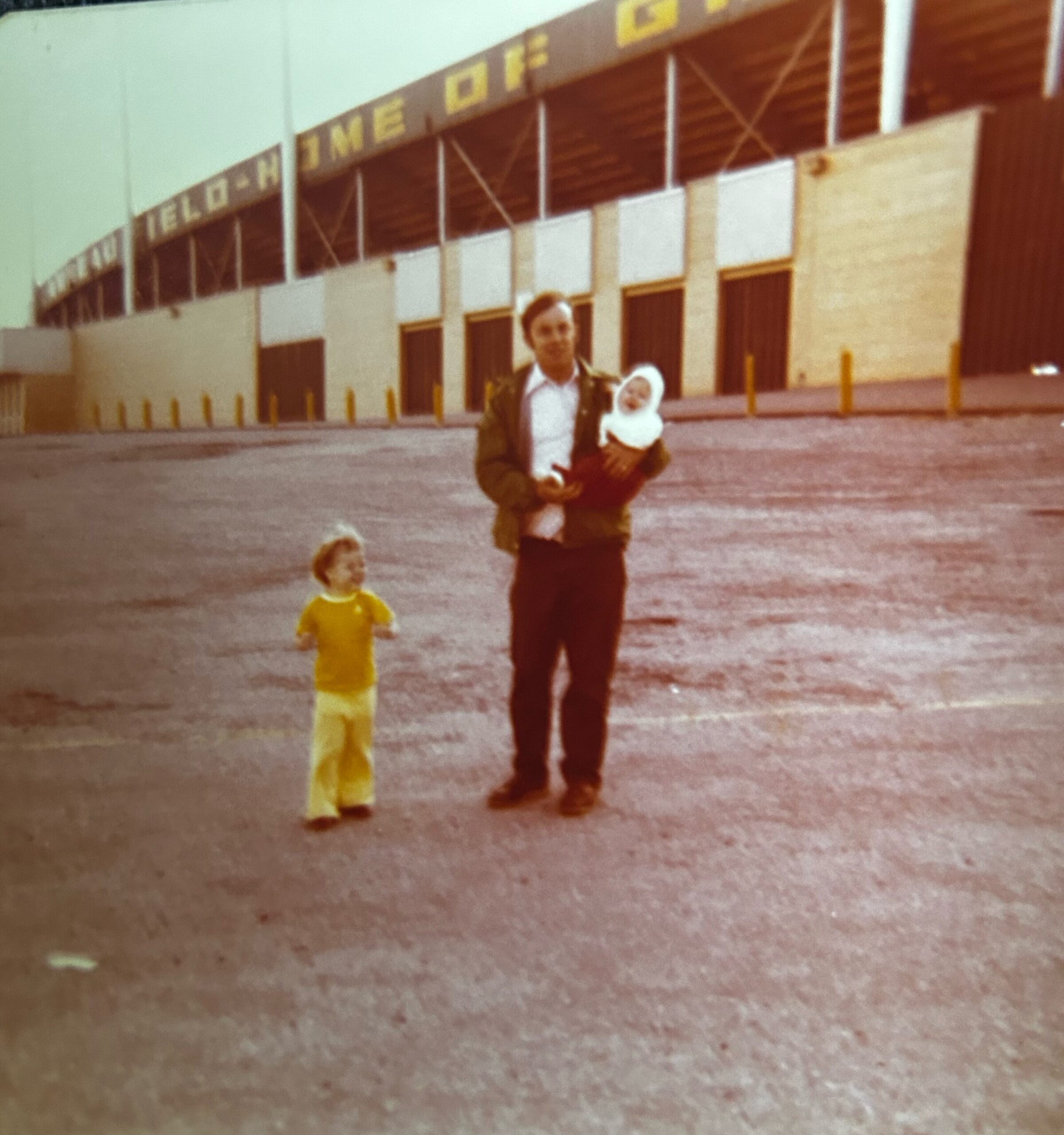 Man in a brown jacket holding a baby, with a young child in yellow standing nearby in a large parking lot, in front of a long building with garage doors.