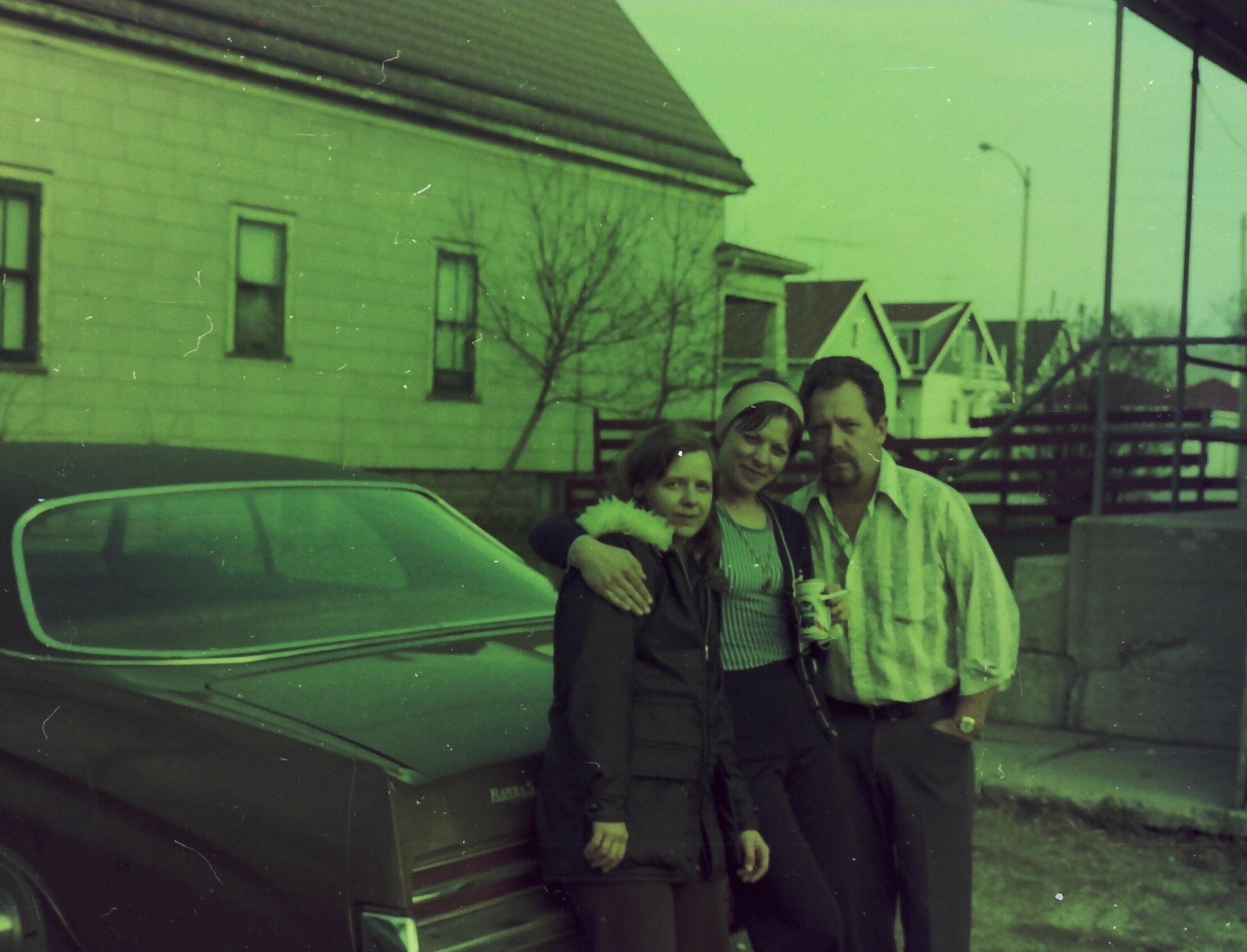 Three people—a man, a woman, and a child—pose together in front of a parked car on a residential street, smiling at the camera.