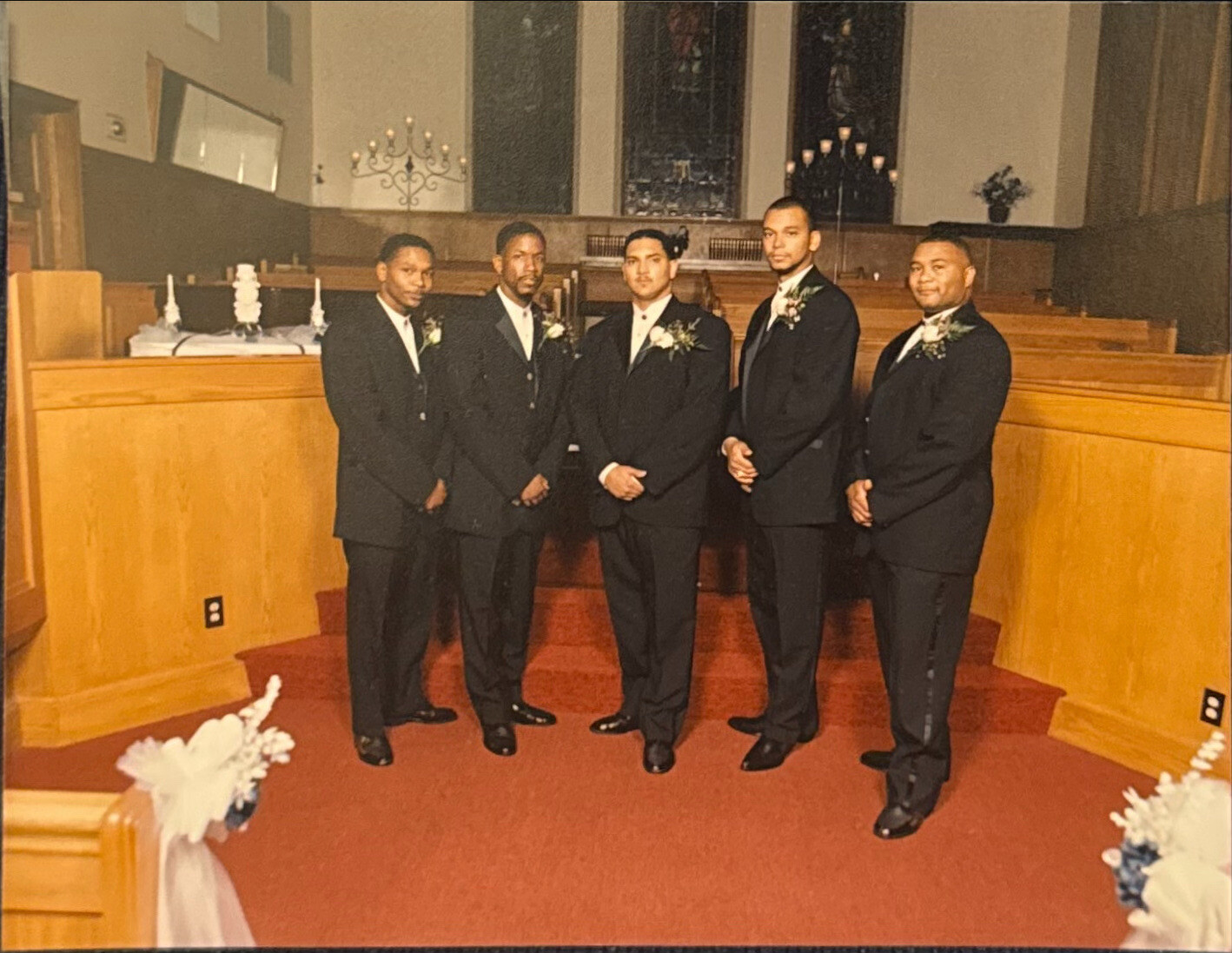 Six men in black tuxedos with boutonnieres pose together in a church aisle for a wedding photo.