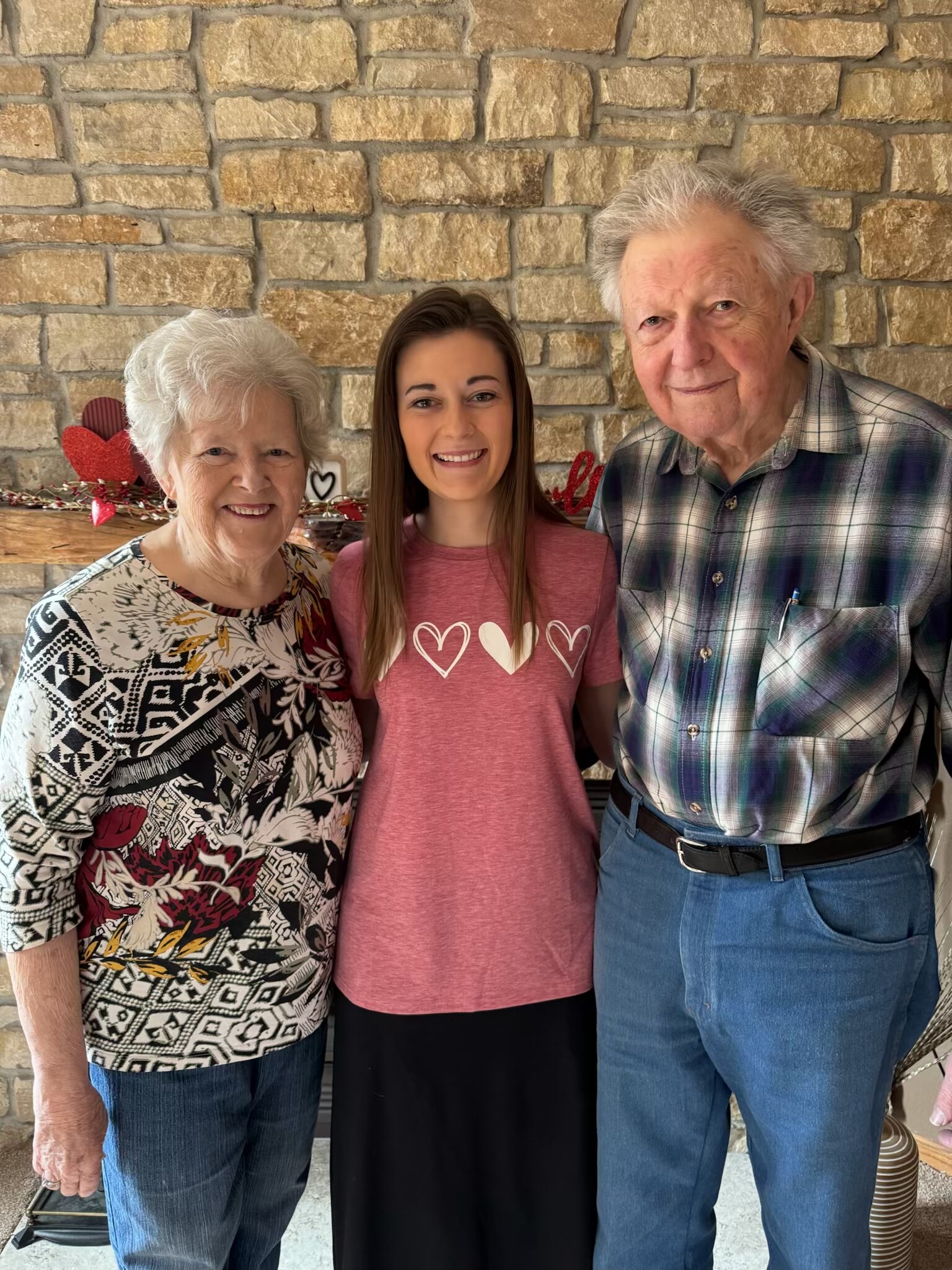 Three generations smiling together: elderly grandmother, young woman, and elderly grandfather in front of a stone wall with Valentine heart decorations on a mantel behind them.