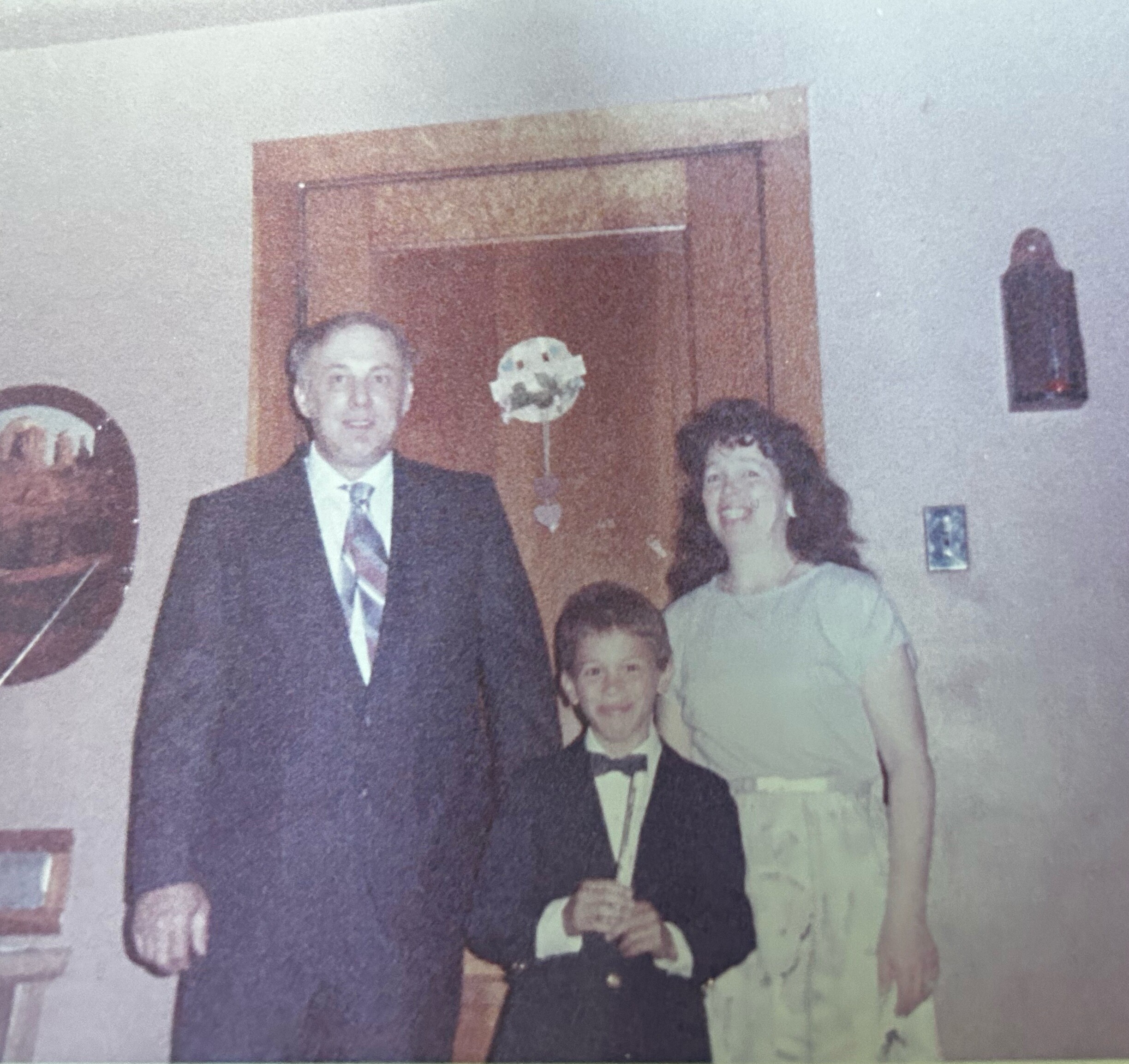 Family photo indoors: man in a suit, woman in a dark dress, and two boys in suits posing together near a wooden door.