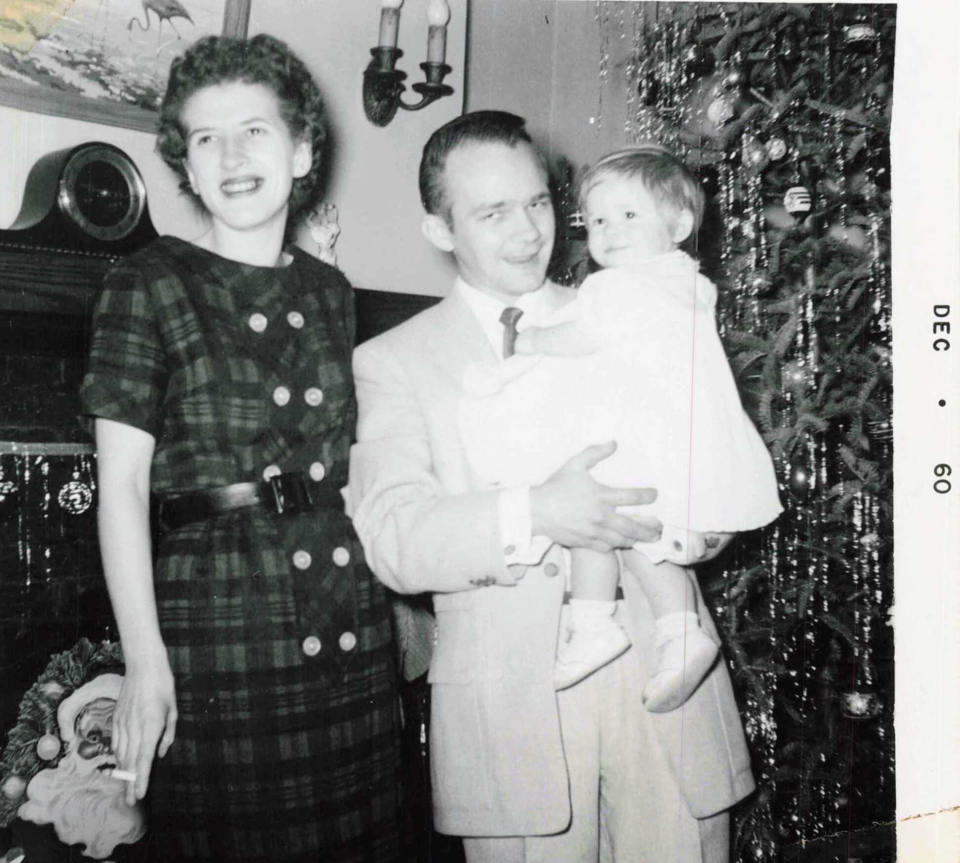 Family posing for a holiday photo: woman in a plaid dress, man in a suit holding a young child by a Christmas tree.