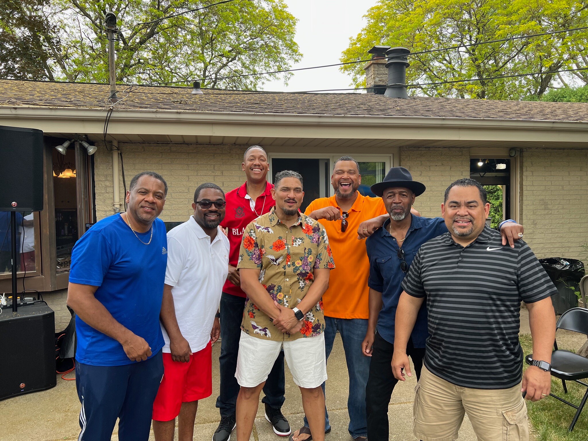 Group of men posing together outdoors in a backyard, smiling and wearing colorful shirts, with a house and trees in the background.