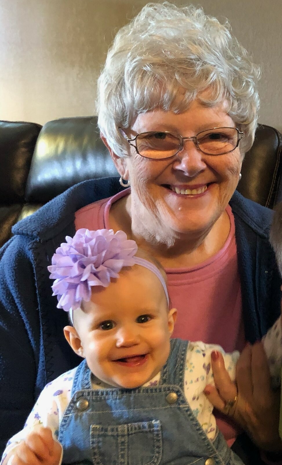 Smiling grandmother with short white hair and glasses sits with a baby girl in denim overalls and a purple flower headband.