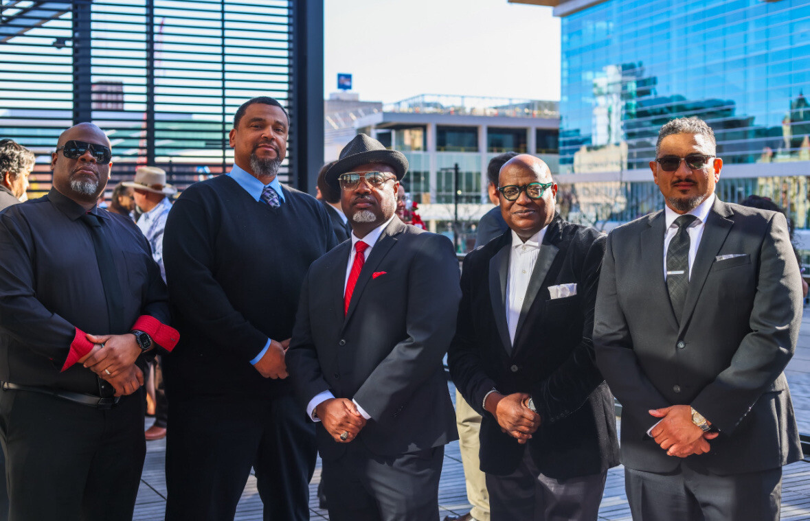 Five men in suits posing together on a rooftop terrace with modern glass buildings in the background.