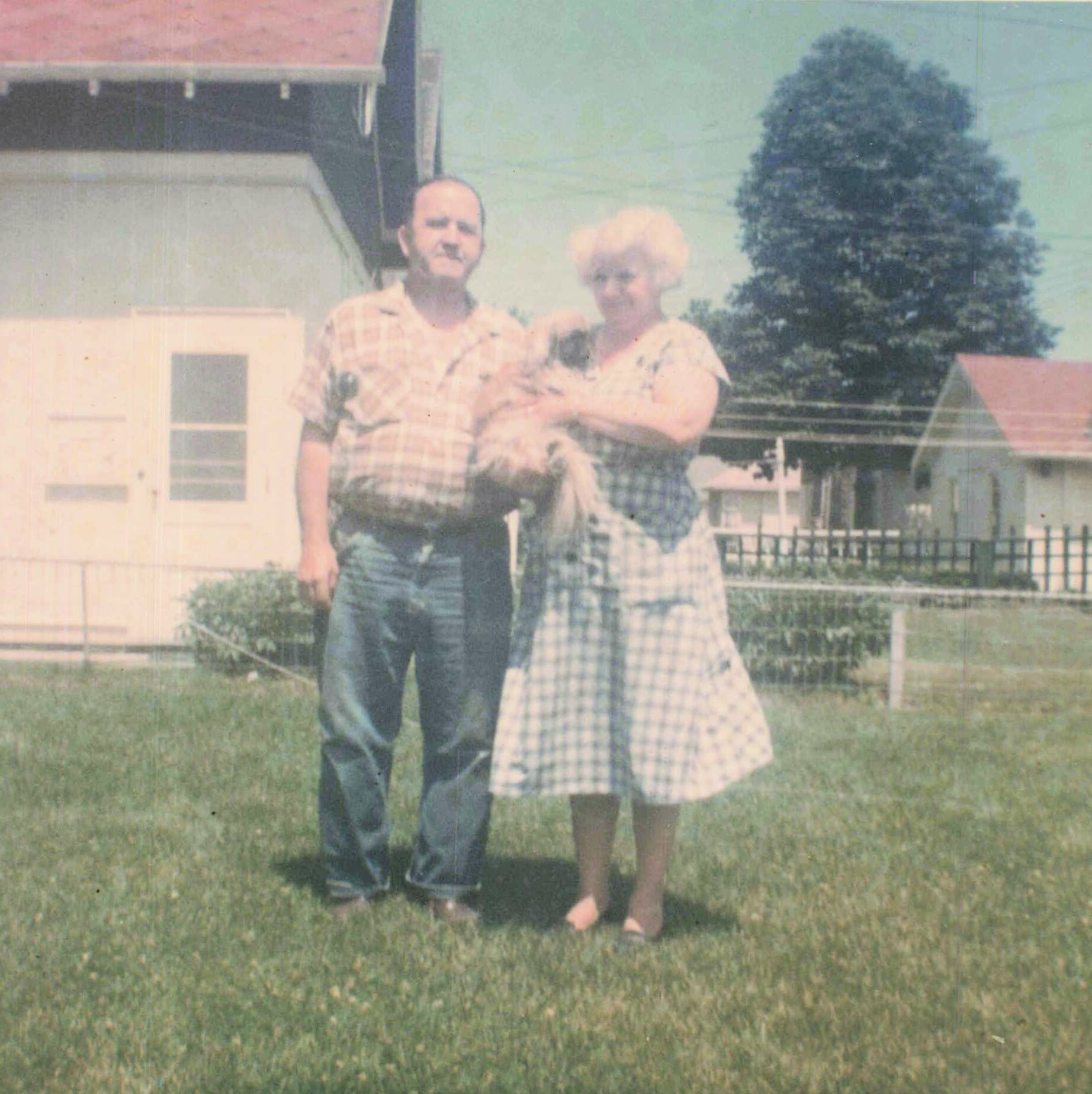 A man and a woman stand together in a front yard, the man in a plaid shirt and jeans, the woman holding a baby between them.