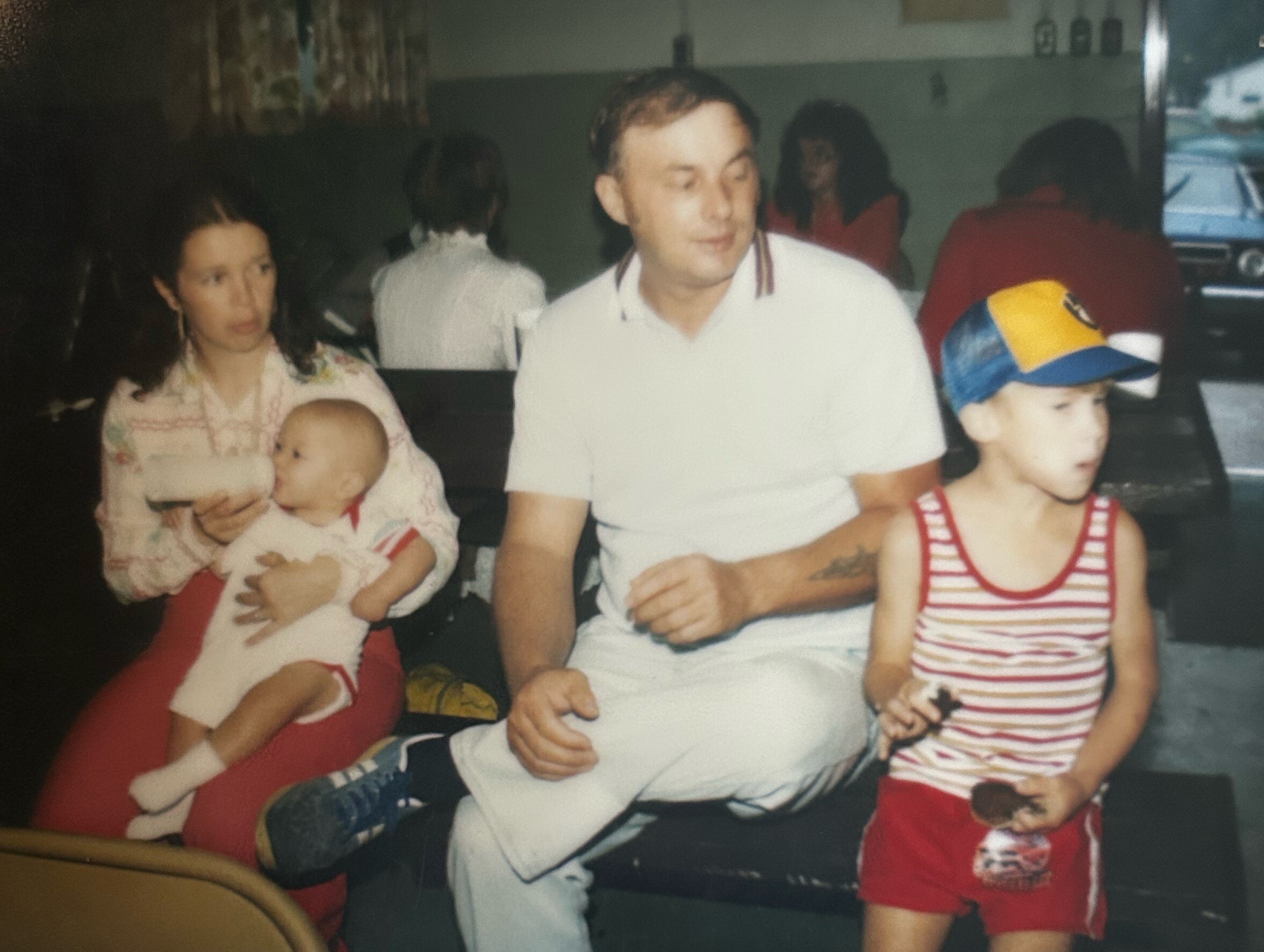 Family at a restaurant table: mother holding a baby, father in a white polo, and a young boy in a striped outfit with a colorful cap on the right.
