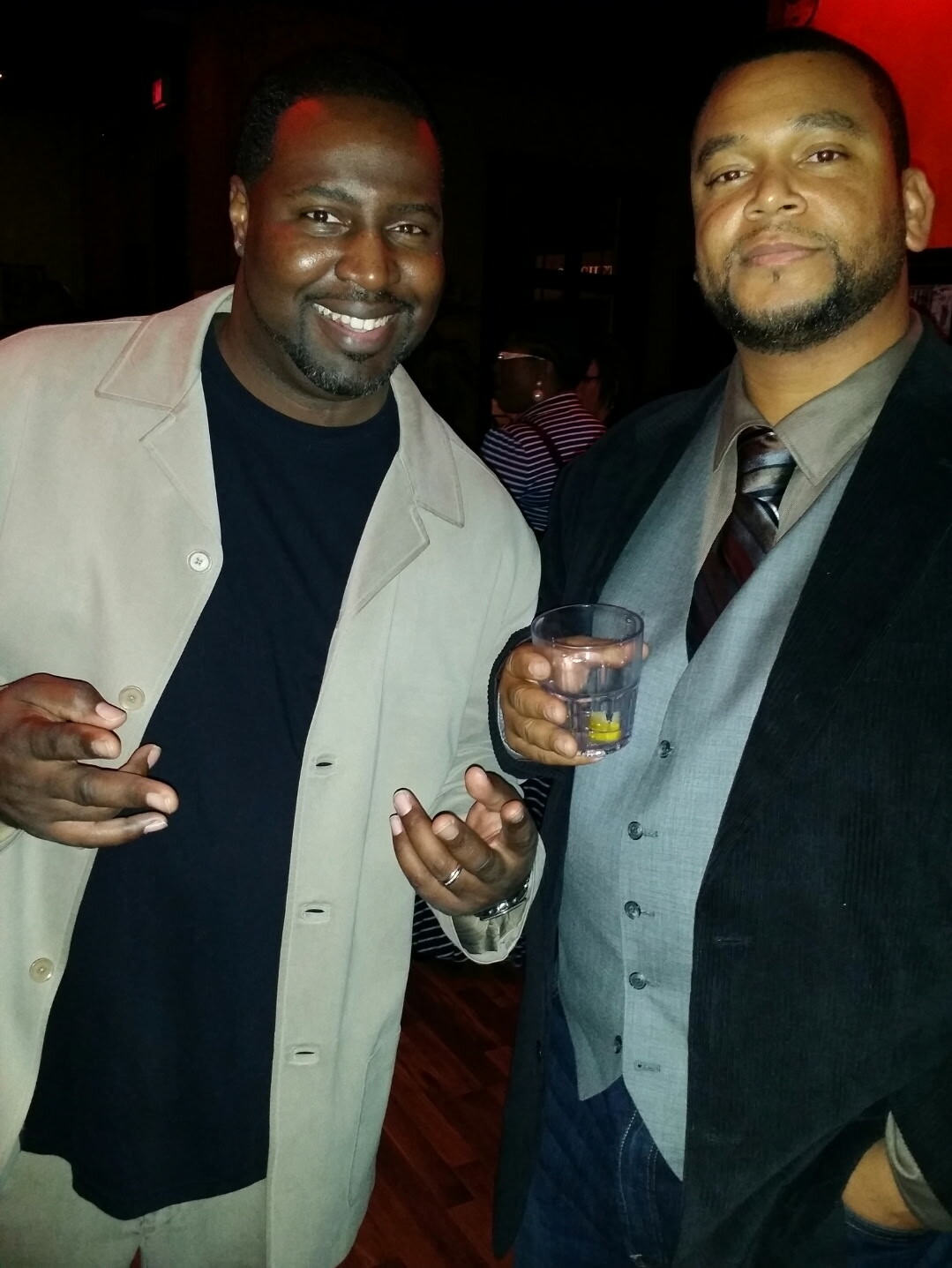 Two men posing and smiling at a dimly lit party; one holds a glass with a lemon slice while the other gestures with his hands.