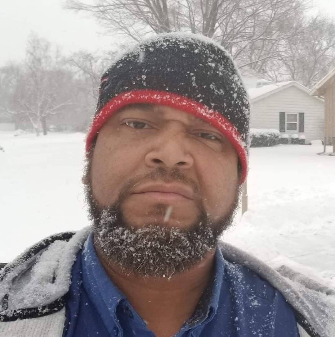 Man outdoors in a snowy yard, wearing a black and red beanie and blue shirt, frost on his beard.