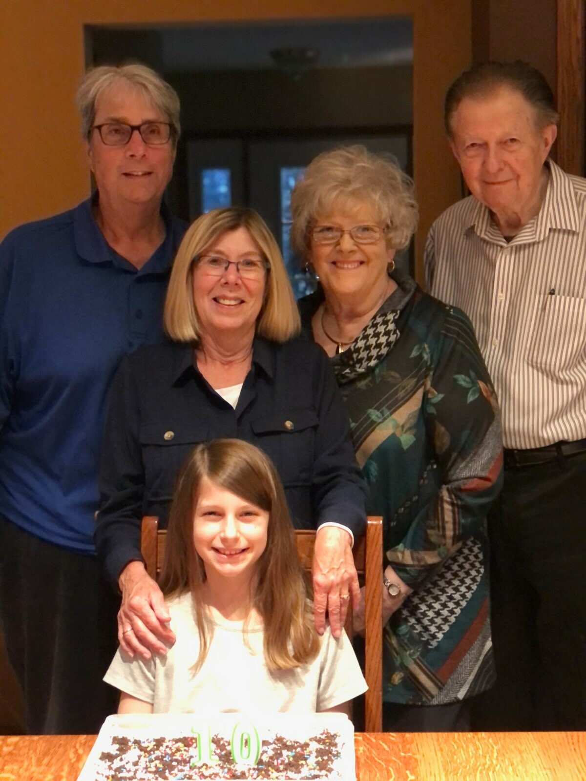 Five family members pose indoors around a table with a birthday cake in front of a smiling girl seated at the table, flanked by four adults standing behind her.