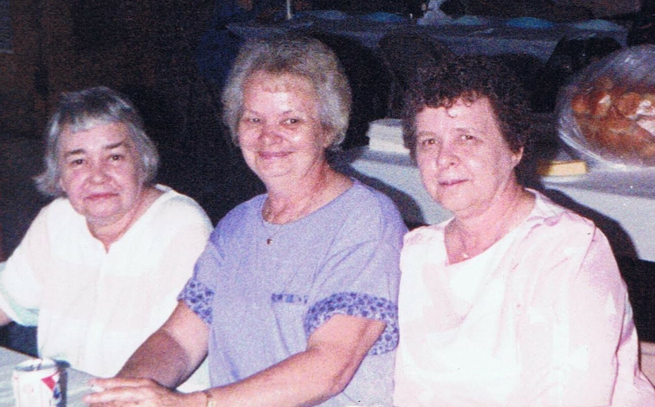 Three elderly women sit together at a table during a meal, smiling at the camera.