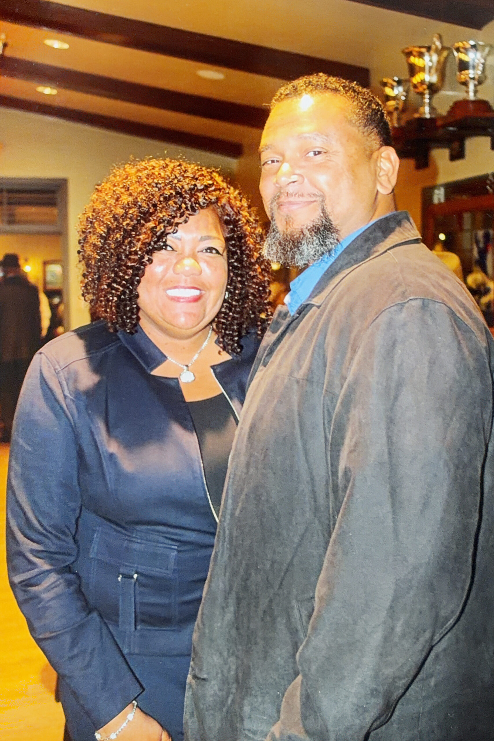 Smiling African American couple posing indoors, woman with curly hair in a dark blazer and necklace, man with blue shirt and brown jacket; trophies on a shelf in the background.