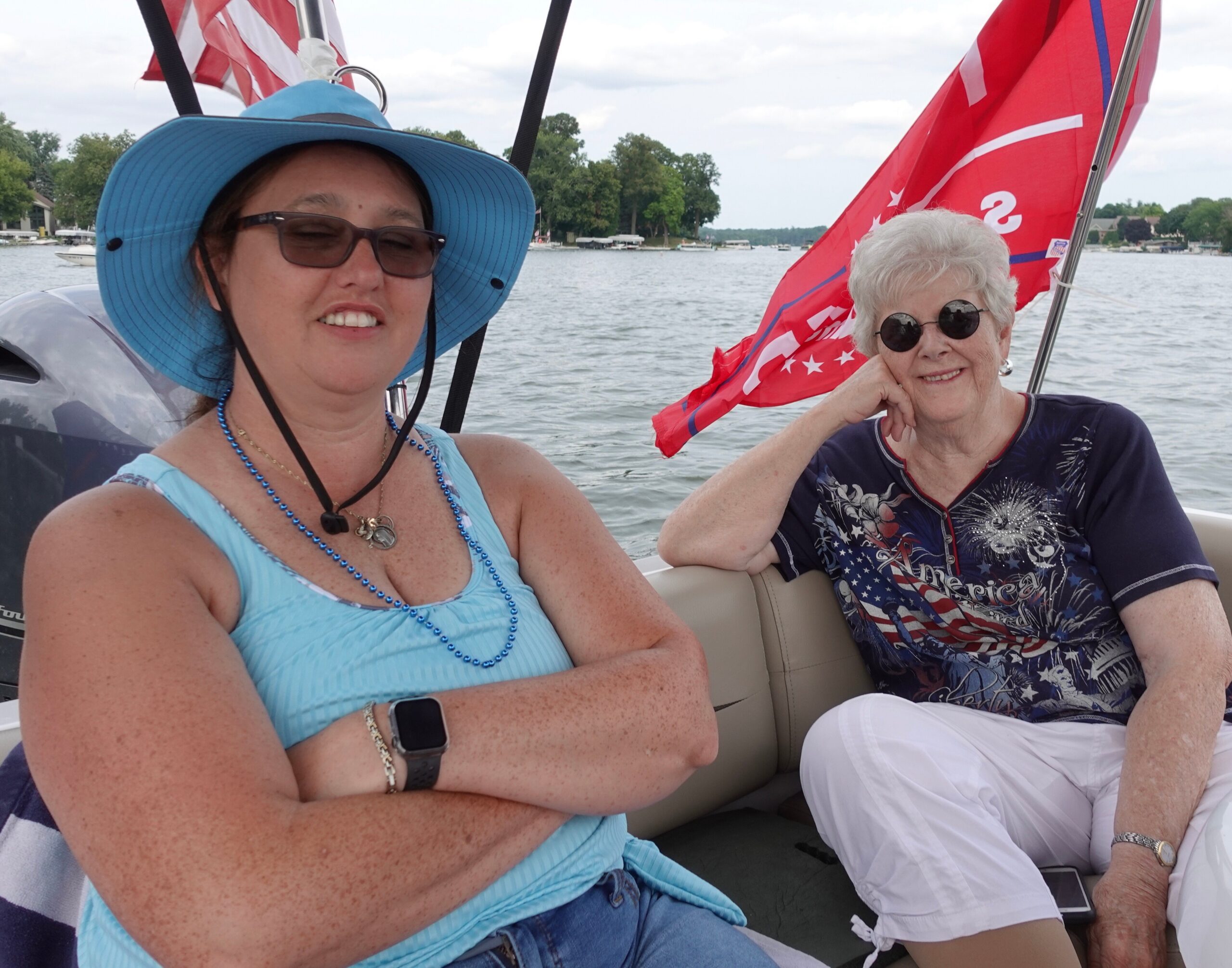 Two women posing on a boat on a lake, one wearing a blue sunhat and sunglasses, the other with short gray hair and round glasses, red flags in the background