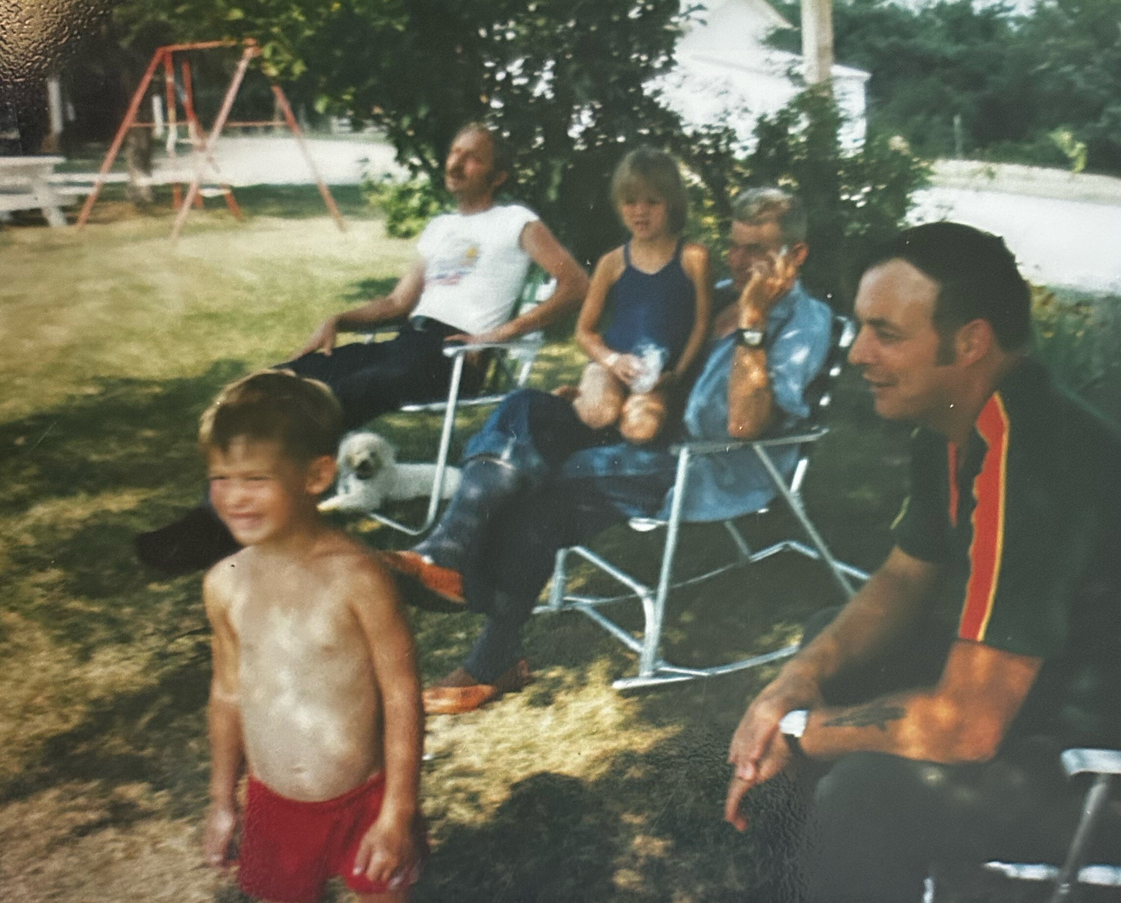 Boy in red swim trunks stands shirtless in a grassy park, with others sitting on folding chairs and a playground visible in the background.