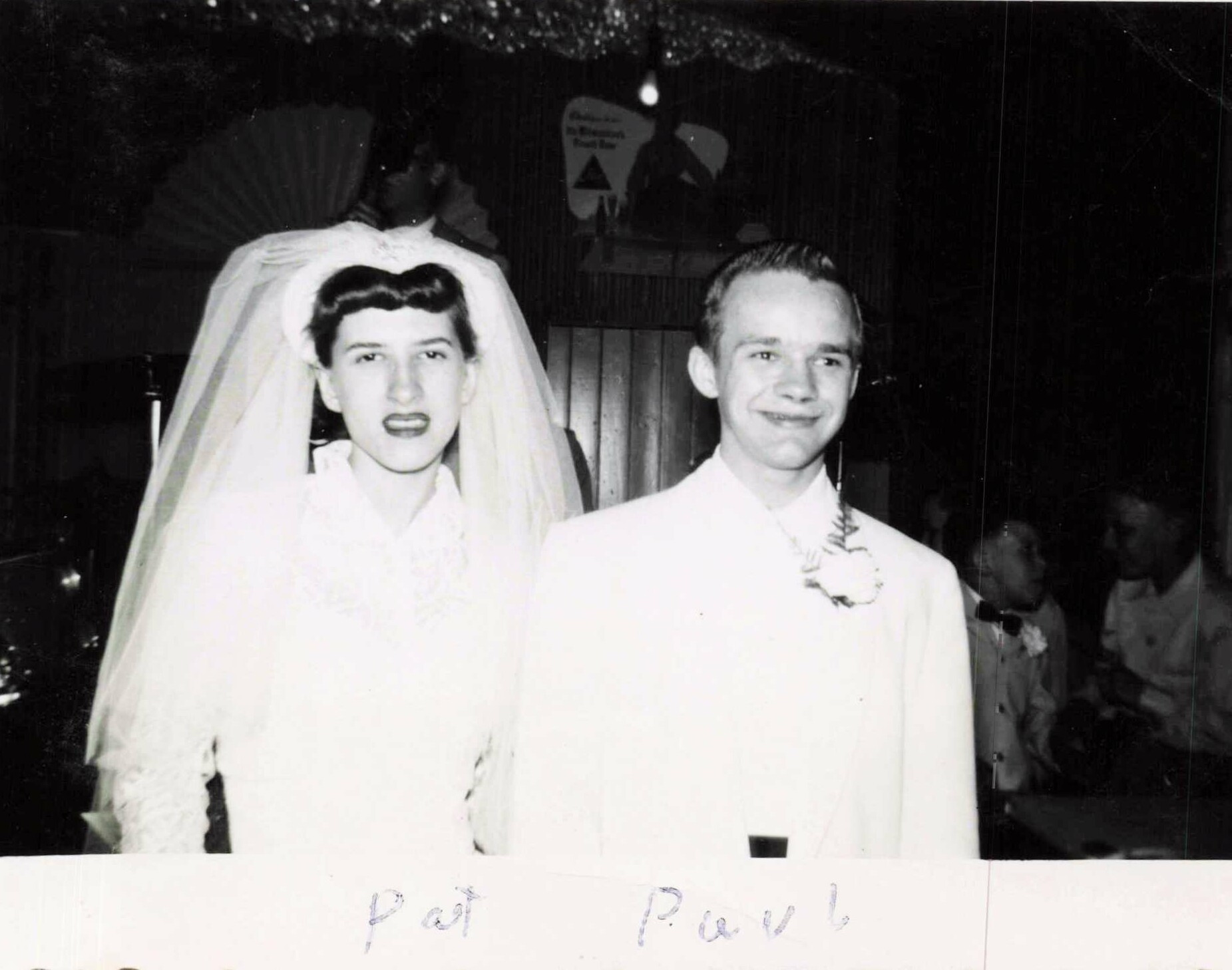 Black-and-white wedding photo of a bride in a veil standing next to a smiling groom in a light suit, with guests in the background