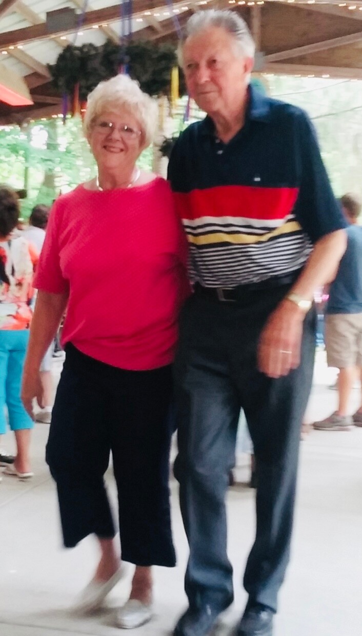 Smiling elderly couple standing arm in arm at an outdoor event; woman in a pink top and black pants, man in a dark polo with red stripe.