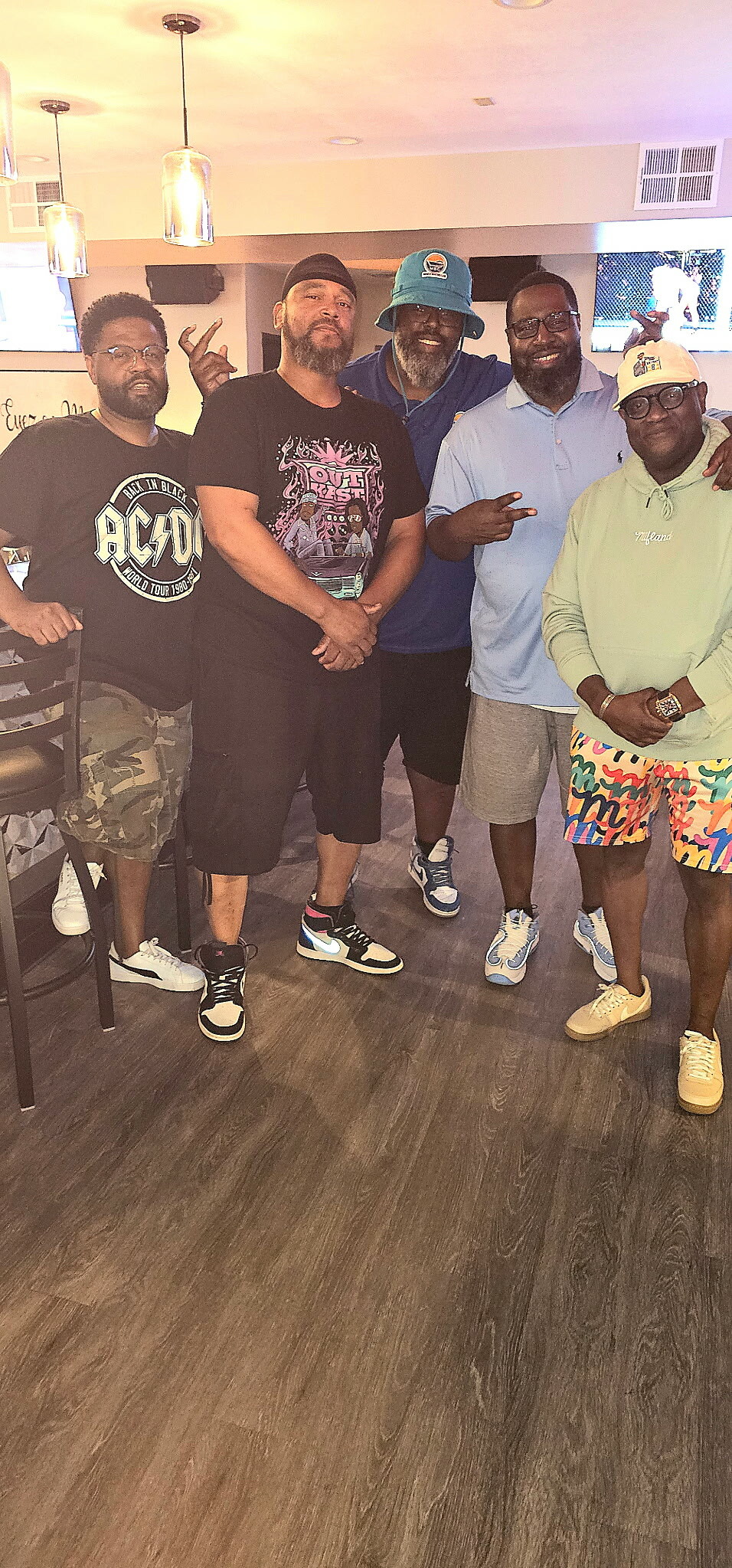 Five men standing together indoors, posing for a group photo with casual outfits and smiles, a bar area and stools visible in the background.