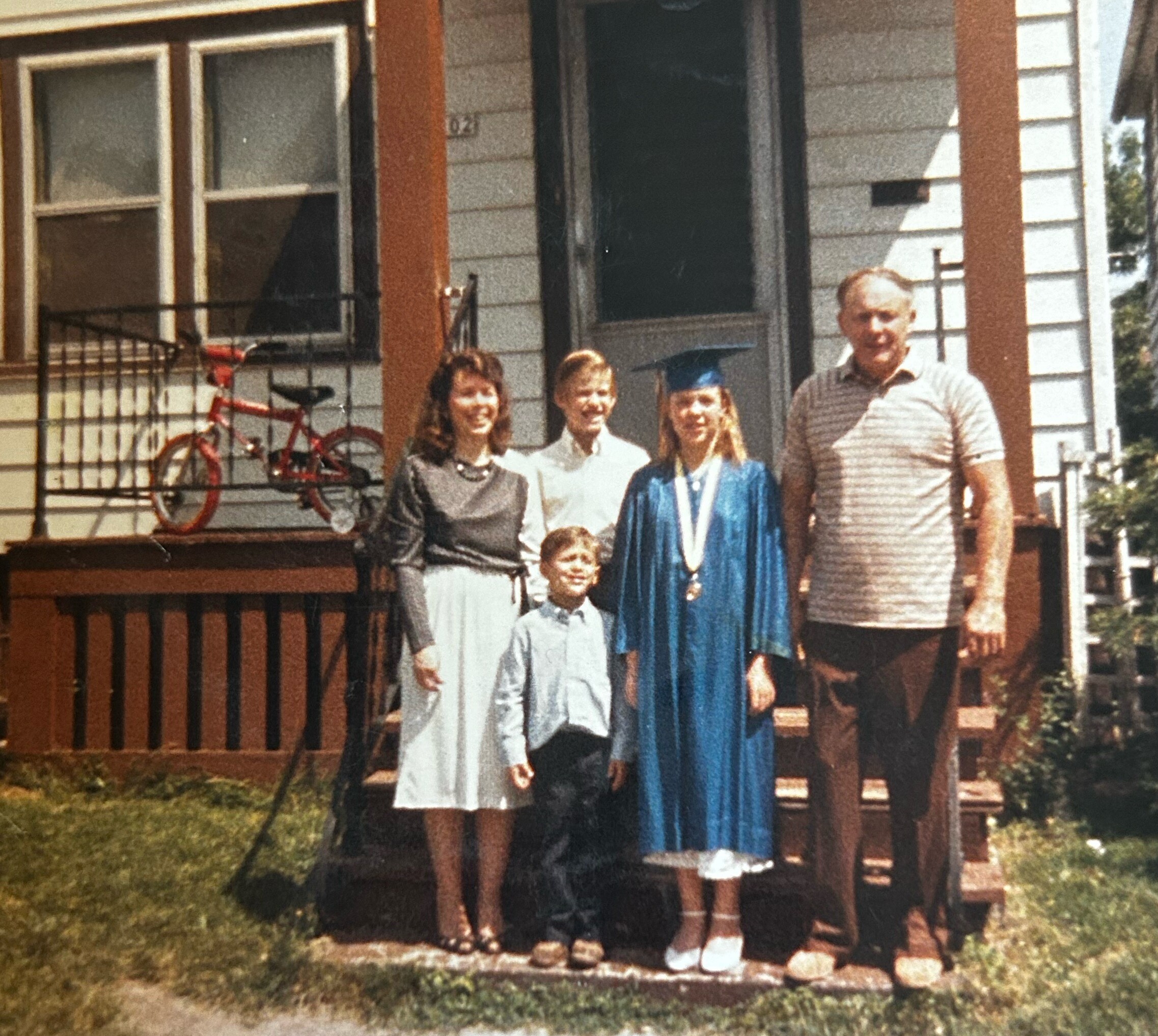 Family group standing on the front porch of a suburban house, with a graduate in a blue gown and cap in the center, plus adults and children wearing casual clothes, and a red bike on the railing behind them.