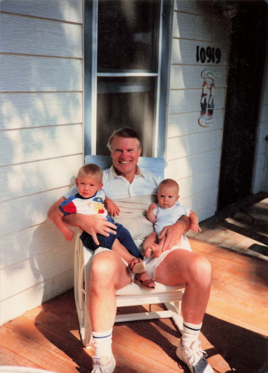 Older man sits in a white chair on a wooden porch, cradling two young children on his lap.