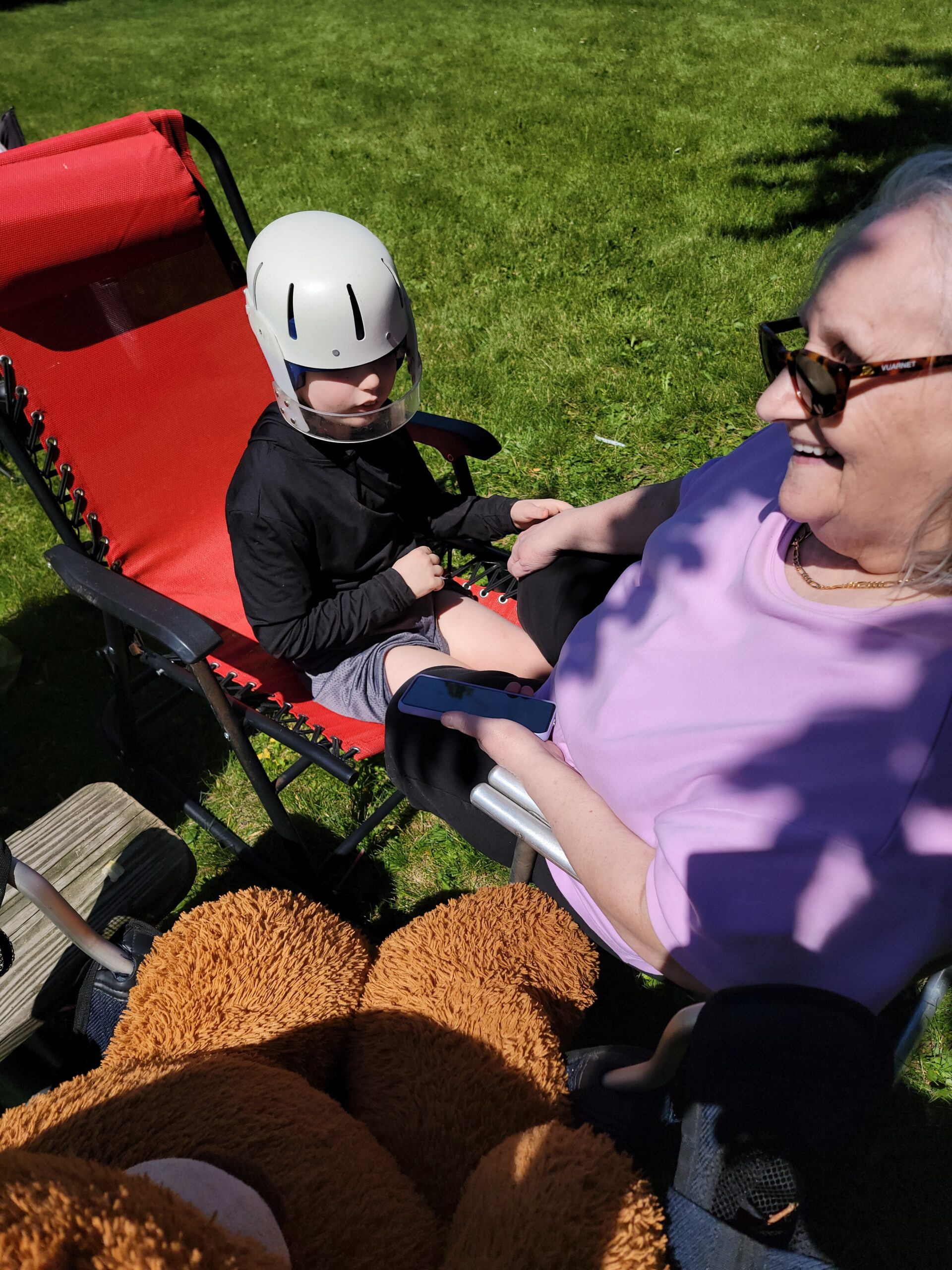 Child wearing a white helmet sits in a red outdoor chair on a sunny grassy lawn, with a smiling older woman beside him holding a phone and wearing sunglasses; brown plush toys are nearby.