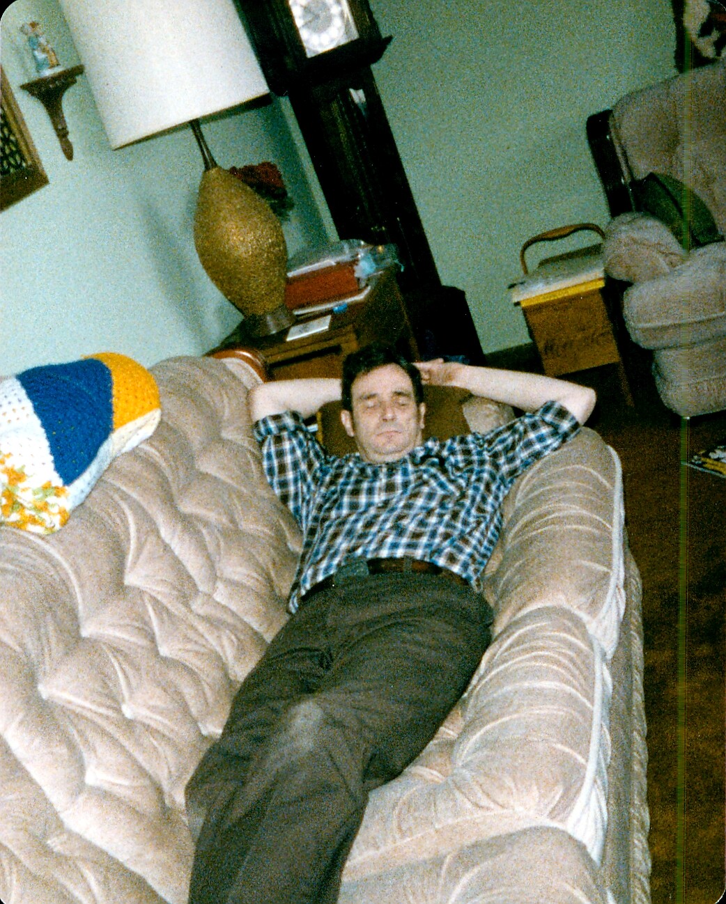 Man resting on a tufted beige sofa in a living room with his hands behind his head, wearing a blue plaid shirt and dark pants.
