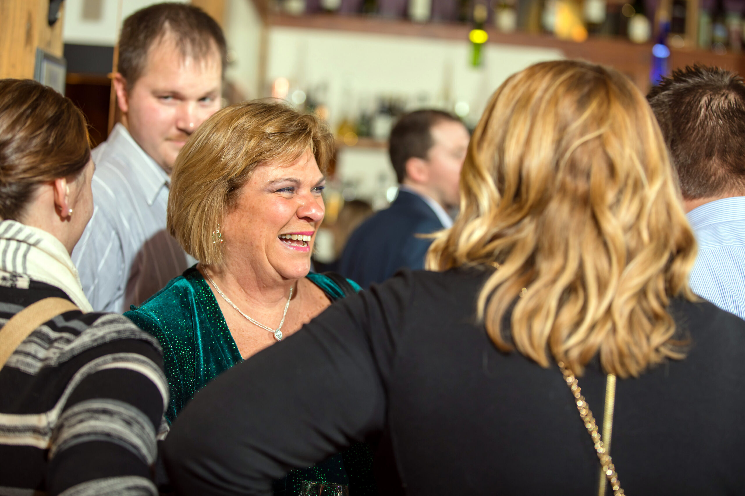 Smiling woman in a teal sequined dress at a crowded social gathering, surrounded by friends.