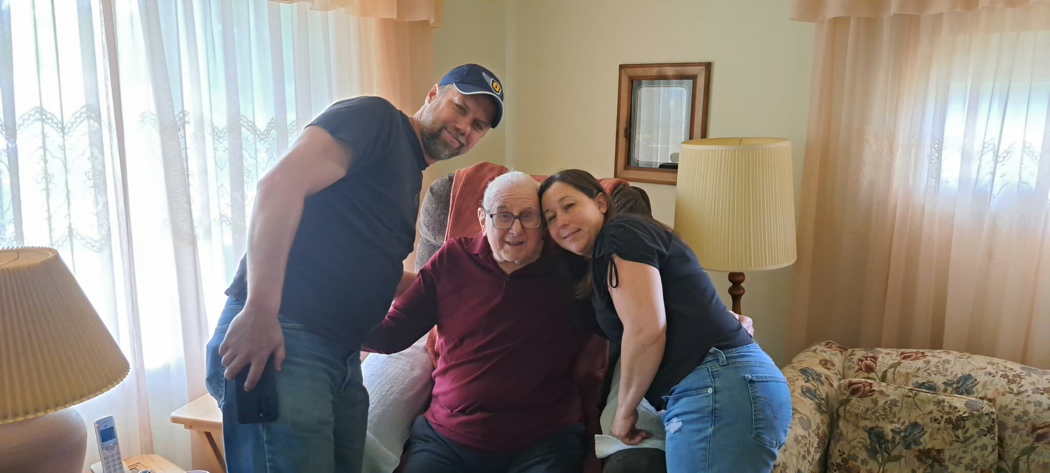Three adults lean in and smile with an elderly man seated in a chair in a cozy living room, posing for a photo.