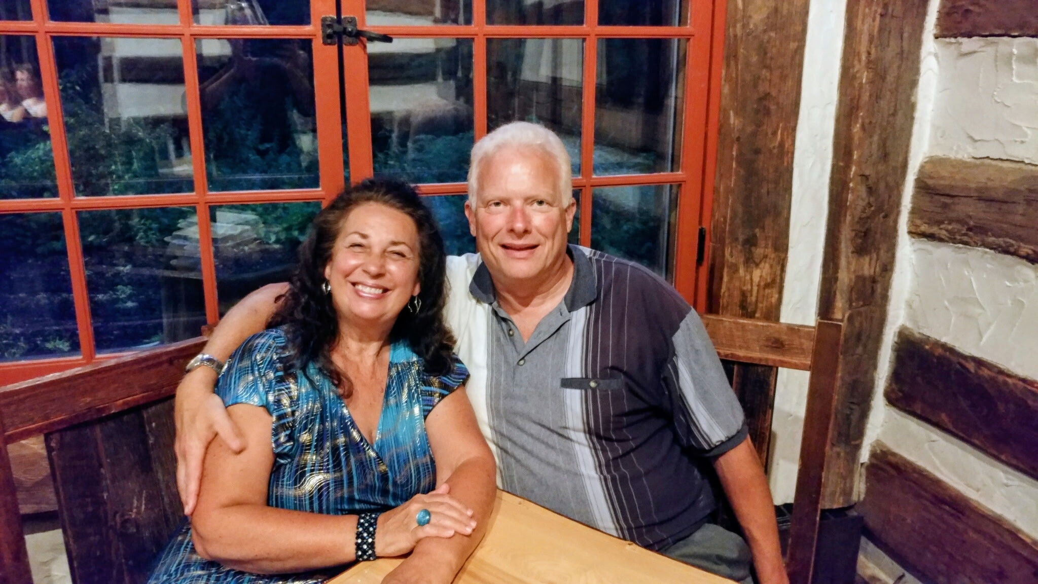 Smiling couple sitting close together at a wooden table in a rustic dining area with red-framed windows behind them.