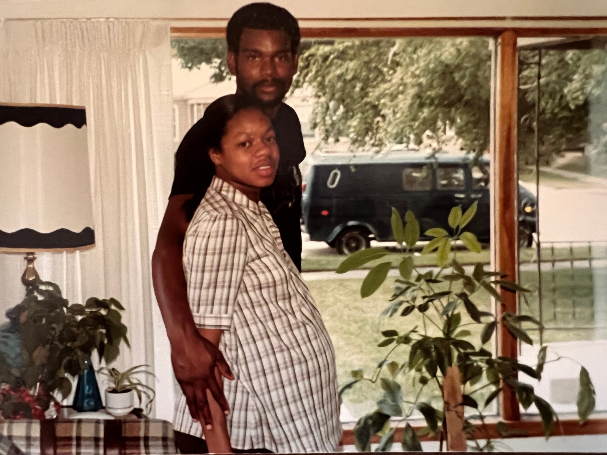 Couple posing together indoors; man stands behind woman with his arm around her shoulders. Stratified home setting visible in background.