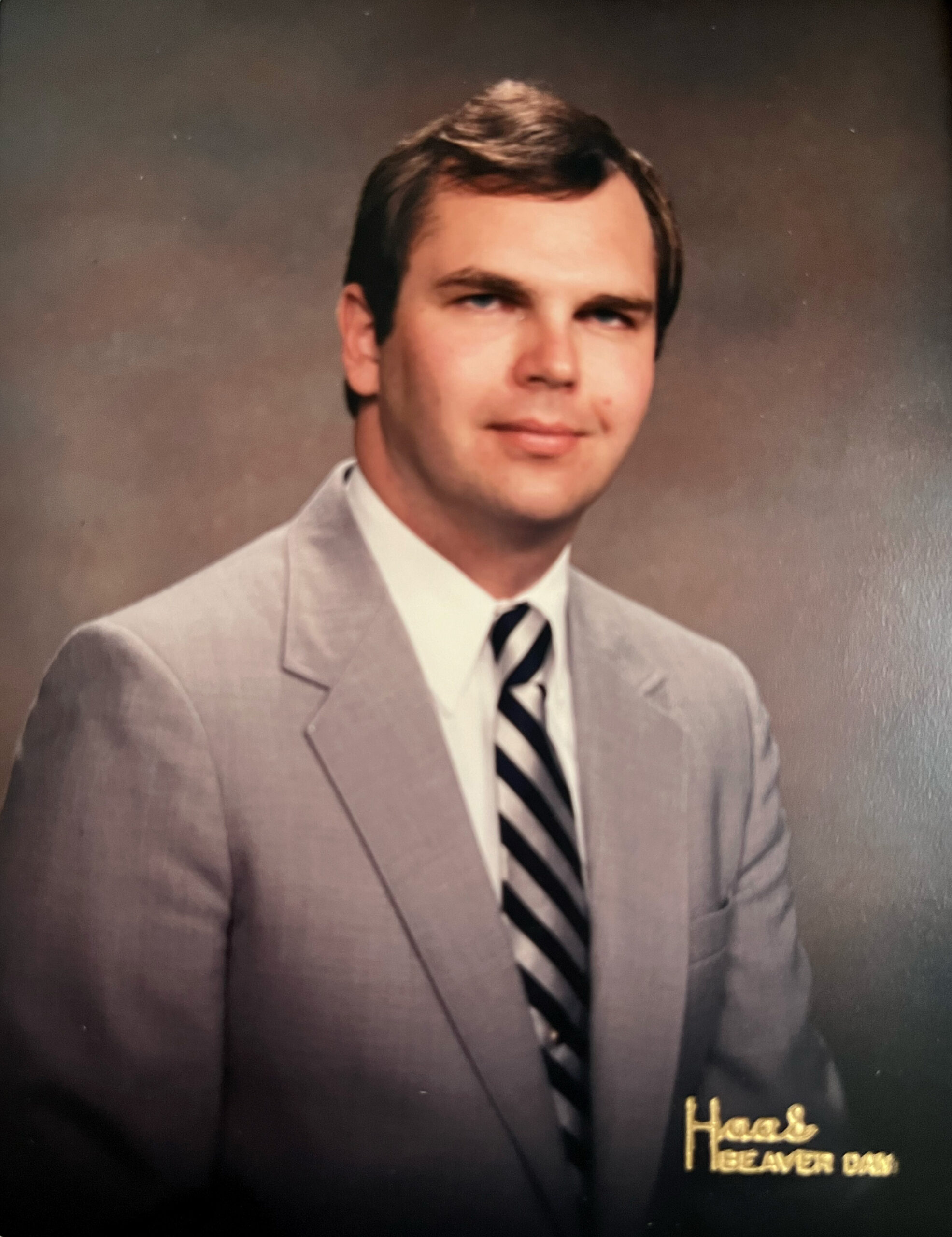 Portrait of a man in a light gray suit and striped tie, posed studio backdrop; gold text appears in the lower right corner, likely part of a yearbook page.