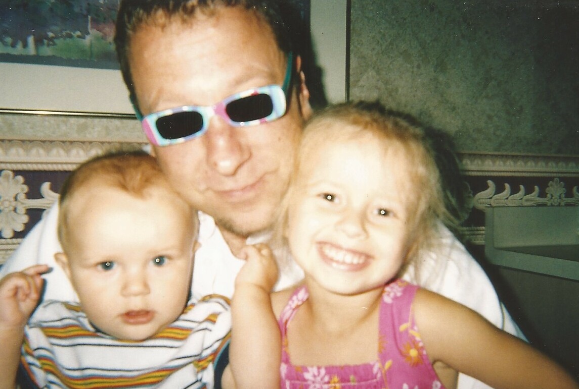 Man wearing colorful sunglasses with a smiling girl in pink and a baby in striped shirt on his lap.