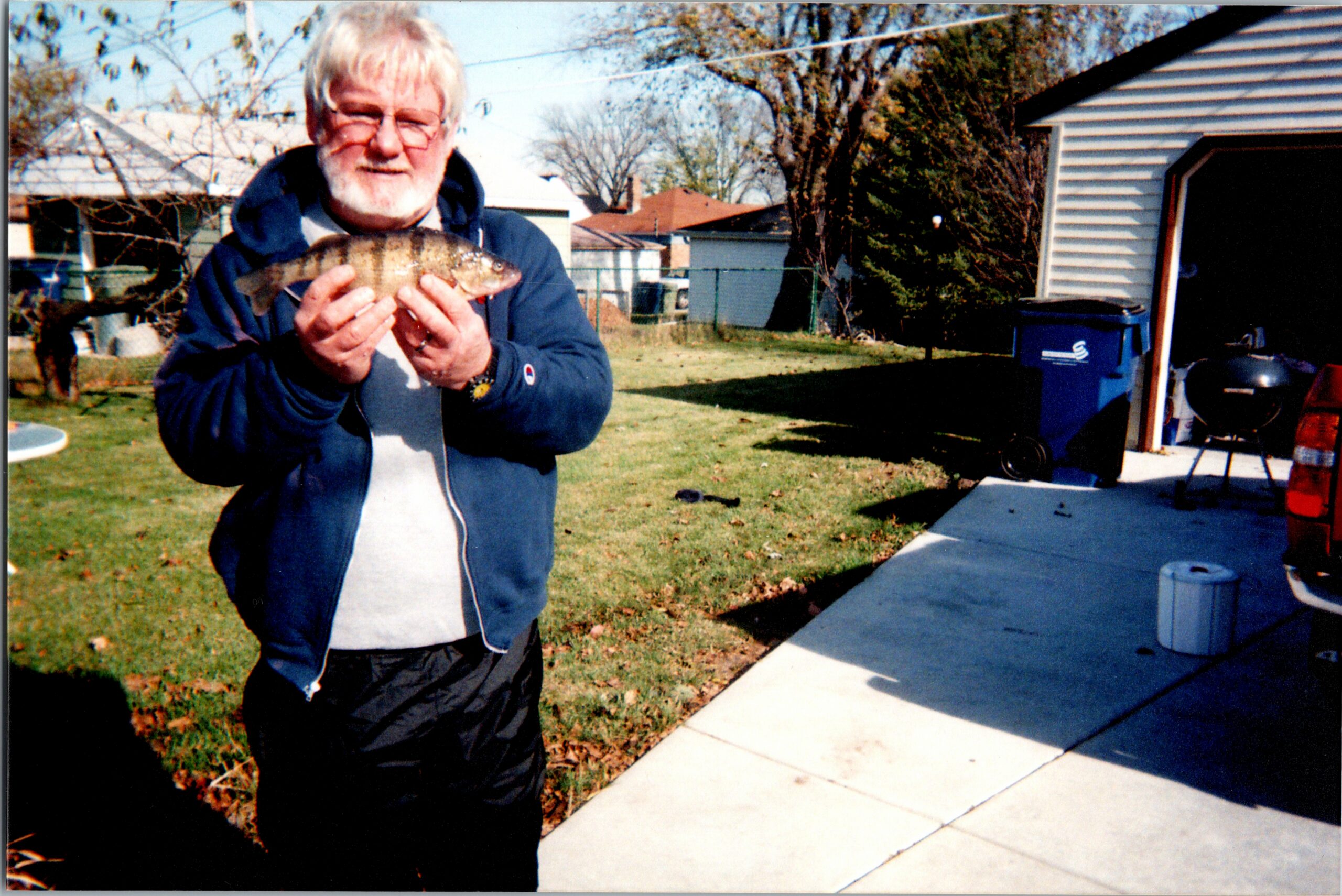 Older man with white beard and glasses holding a striped fish toward the camera in a sunny backyard yard.