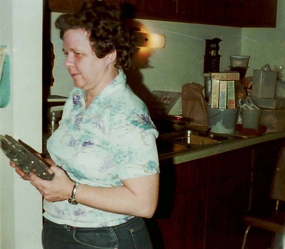 Side-profile of a woman in a floral blouse standing in a cluttered kitchen, holding a dark rectangular tray or plate.