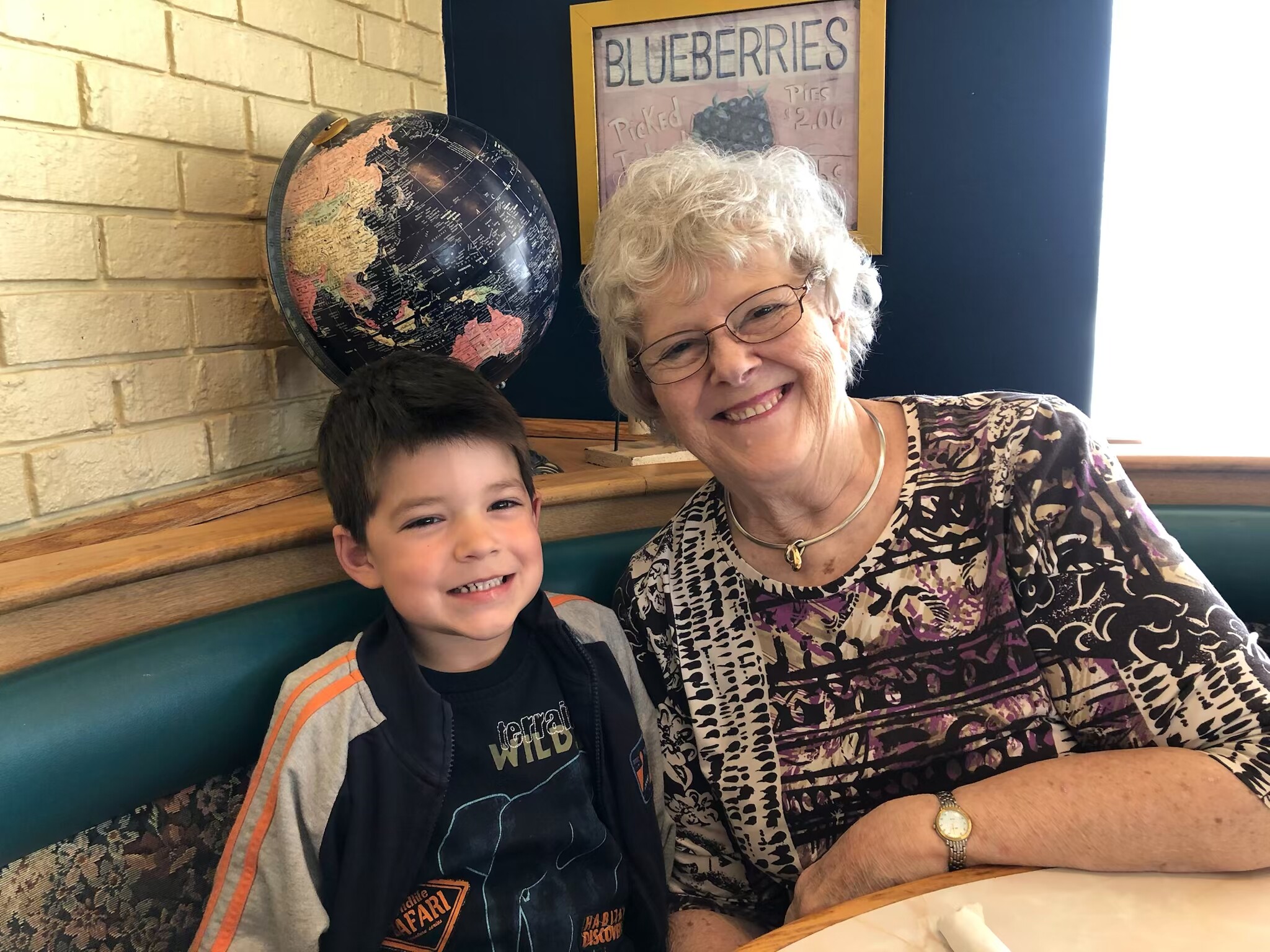 Grandmother and young boy smile for the camera at a cafe booth; a globe sits on the shelf behind them.