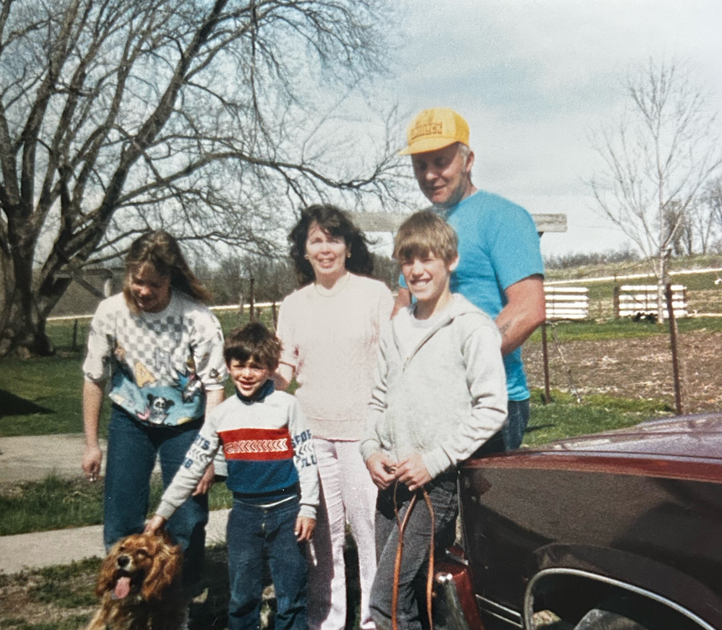 Family group outdoors with a brown dog; man in a yellow cap and blue shirt stands behind children by a wooden fence.