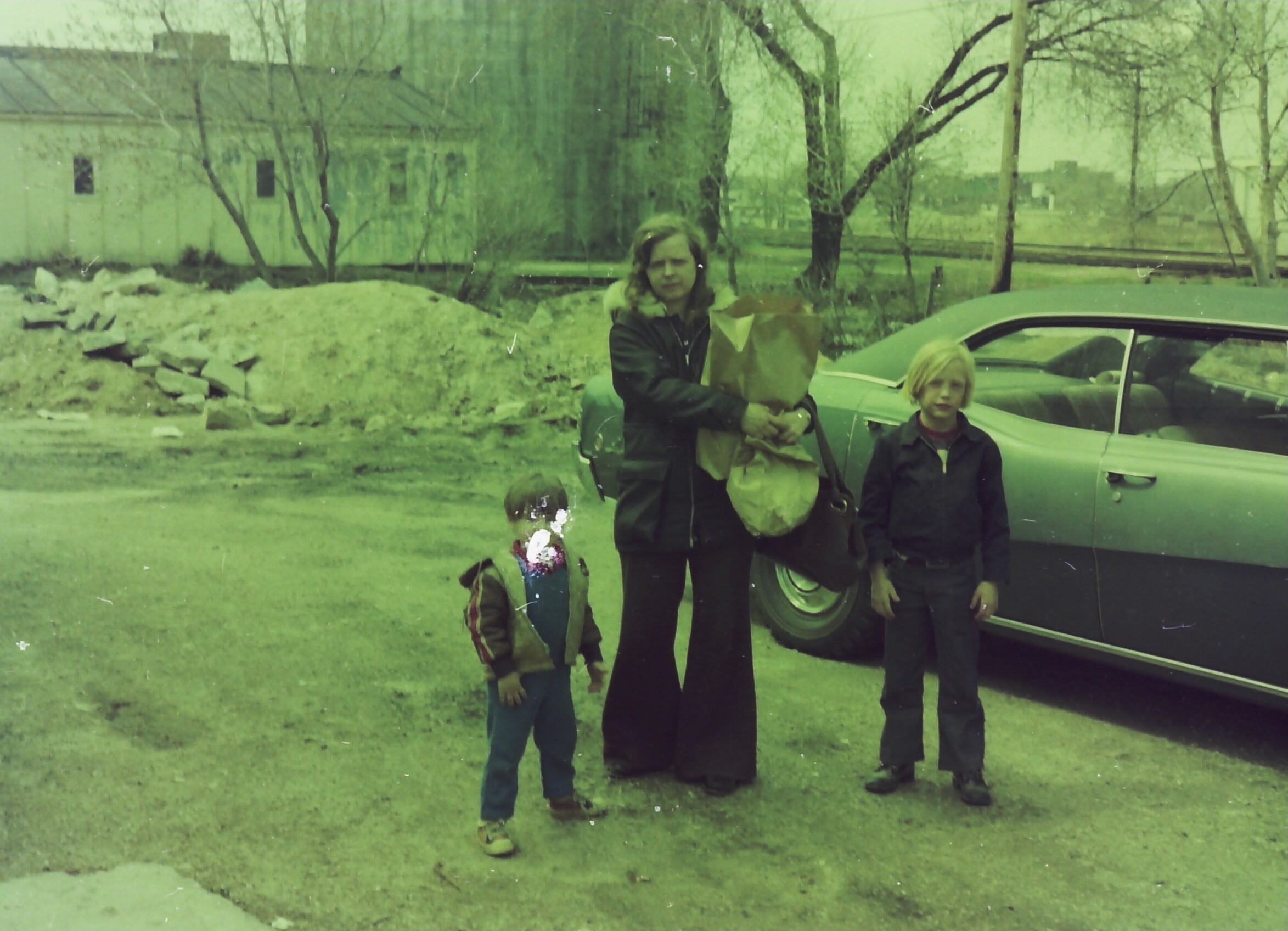 Family of three posing outside near a parked car, with the mother holding a bouquet of flowers and two children nearby in a grassy yard.