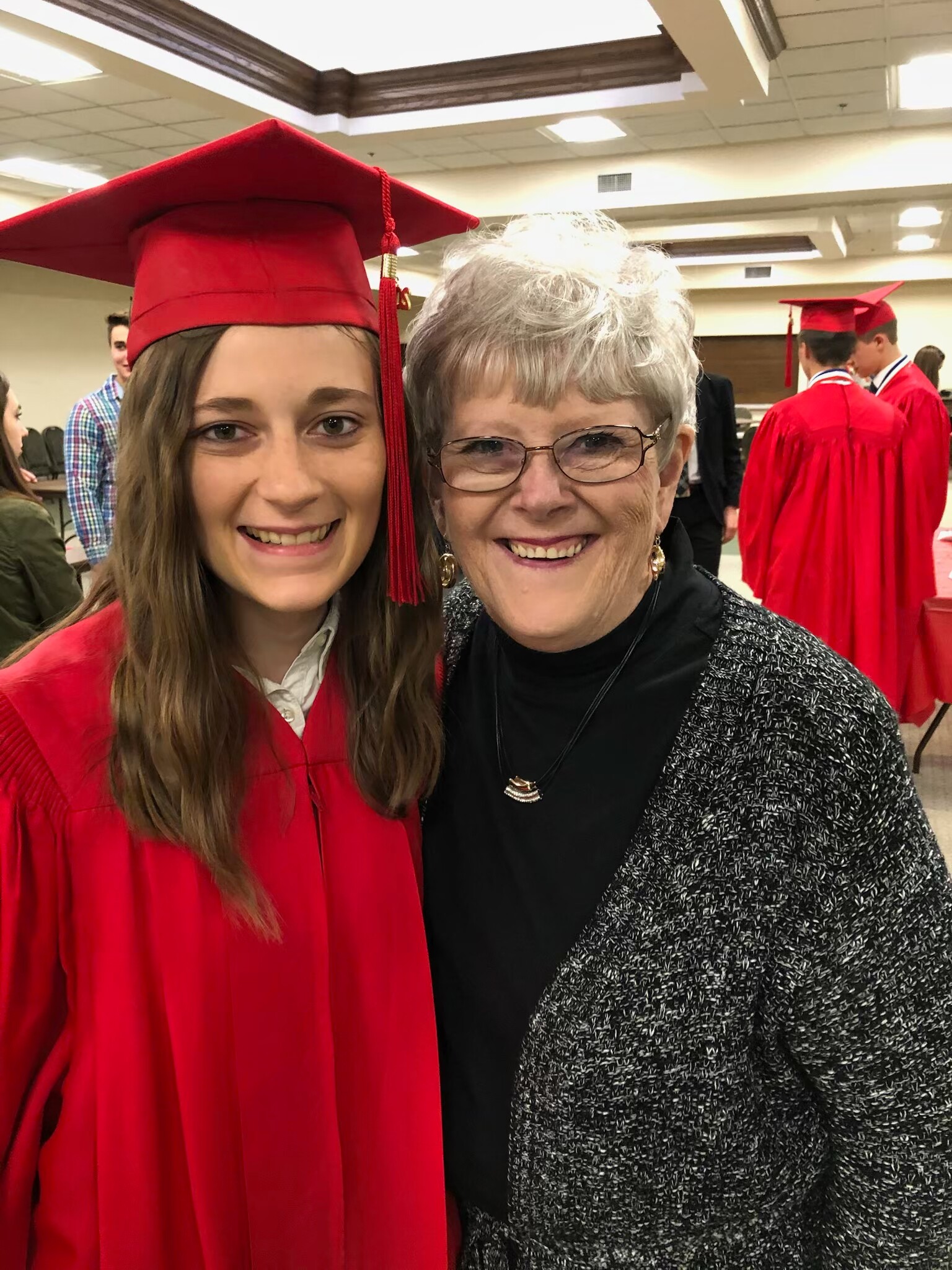 Smiling graduate in a red cap and gown posing with an older woman at a graduation ceremony.