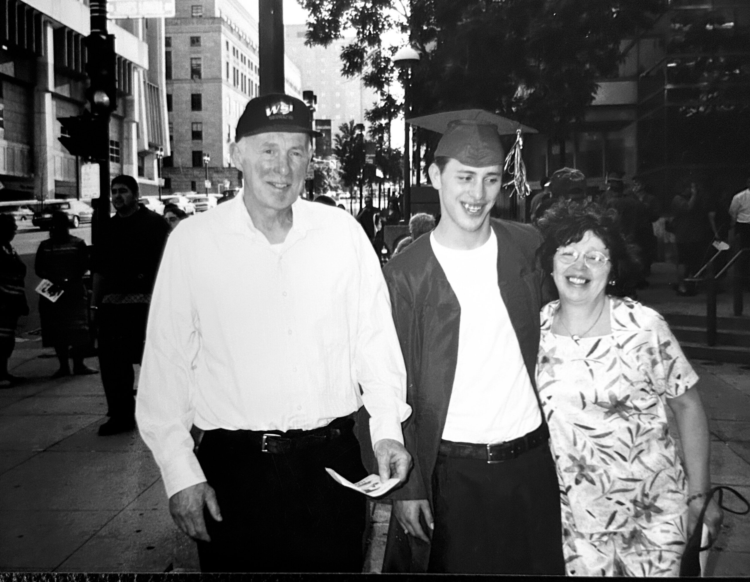 Graduate wearing cap and gown walking with an older man and woman on a city sidewalk, smiling amid a street scene at a graduation celebration.